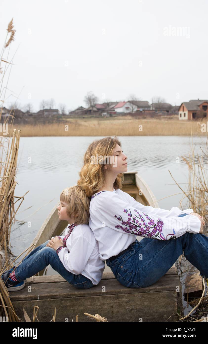 little boy and a young beautiful woman in embroidered national clothes are sitting back to back on the shore of the lake. Family, refugees, unity, sup Stock Photo