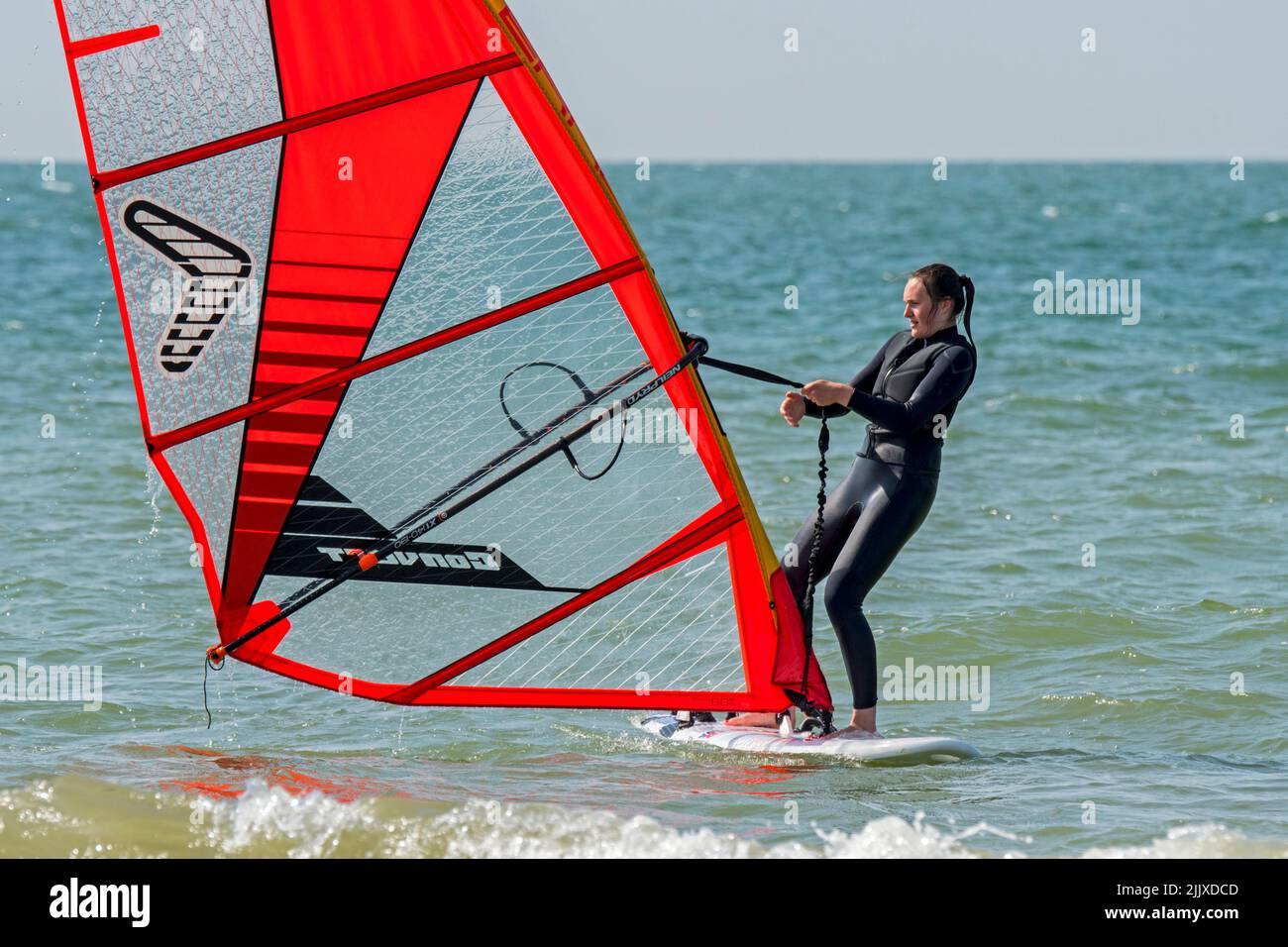 Girl / female recreational windsurfer in black wetsuit practising classic windsurfing and lifting sail at sea Stock Photo