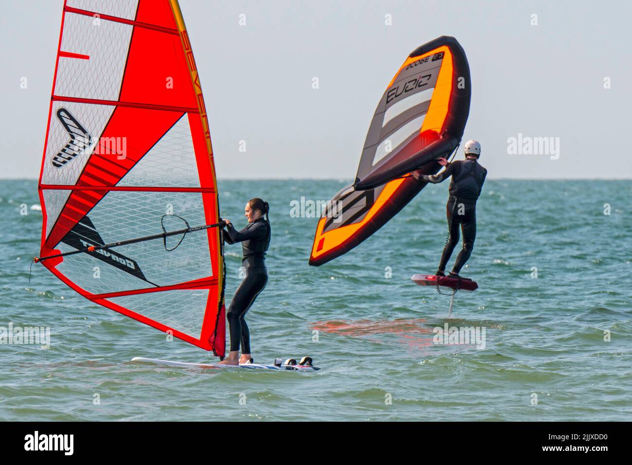 Girl / female recreational windsurfer in black wetsuit practising classic windsurfing and wingboarder / wing boarder wing foiling at sea Stock Photo
