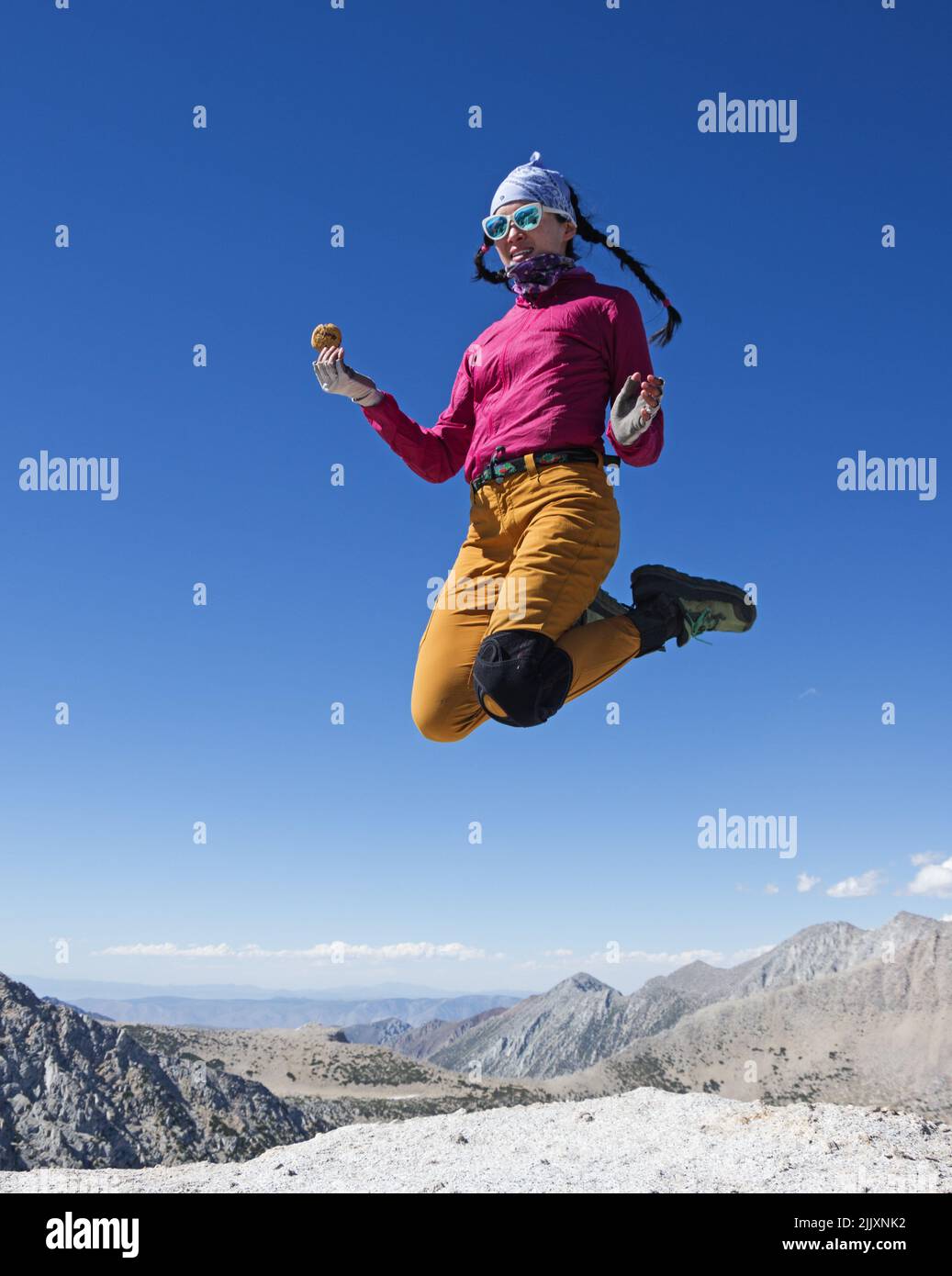 Asian Woman in Sunglasses with a knee brace jumps on top of Mount Hopkins in the Sierra Nevada with a cookie in her hand Stock Photo