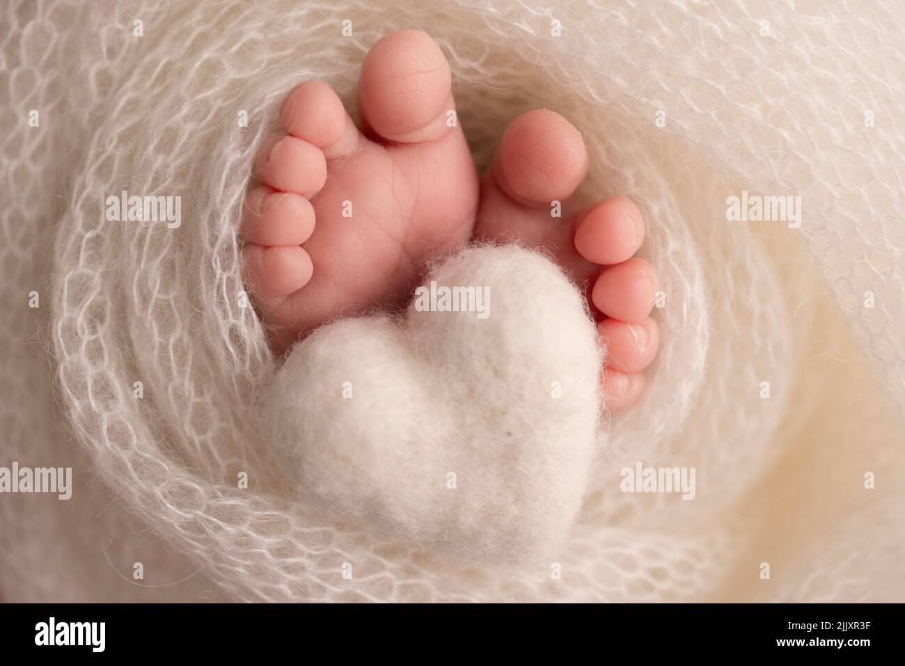 Knitted white heart in the legs of a baby. Soft feet of a new born in a white wool blanket.  Stock Photo
