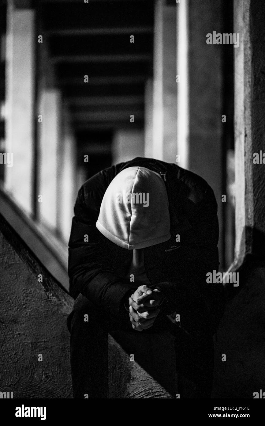A grayscale shot of a man, head down wearing a hood and sitting in an ...