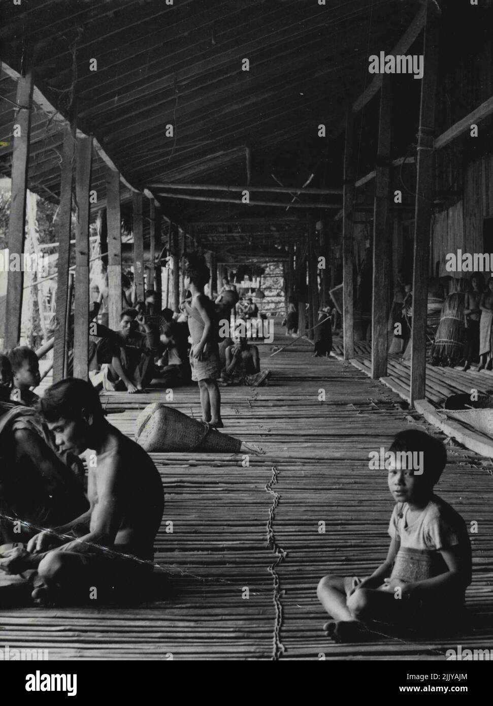 Land Dayaks Of Borneo A Dayak Longhouse: The long communal verandah, with the rooms of the individual households leading off from it. Women are pounding rice, and beside the wooden pounders lie winnowing baskets. Groups of men are gossiping or working, a boy is minding his baby brother, and against the wall are bundles of rattan vine for making mats and ropes. Fifty families, comprising up to 400 people, live in houses opening off this long communal veranda where women pound their rice. Rattan for making ropes is piled against the wall. March 8, 1952. (Photo by W.R. Geddes, Camera Press). Stock Photo