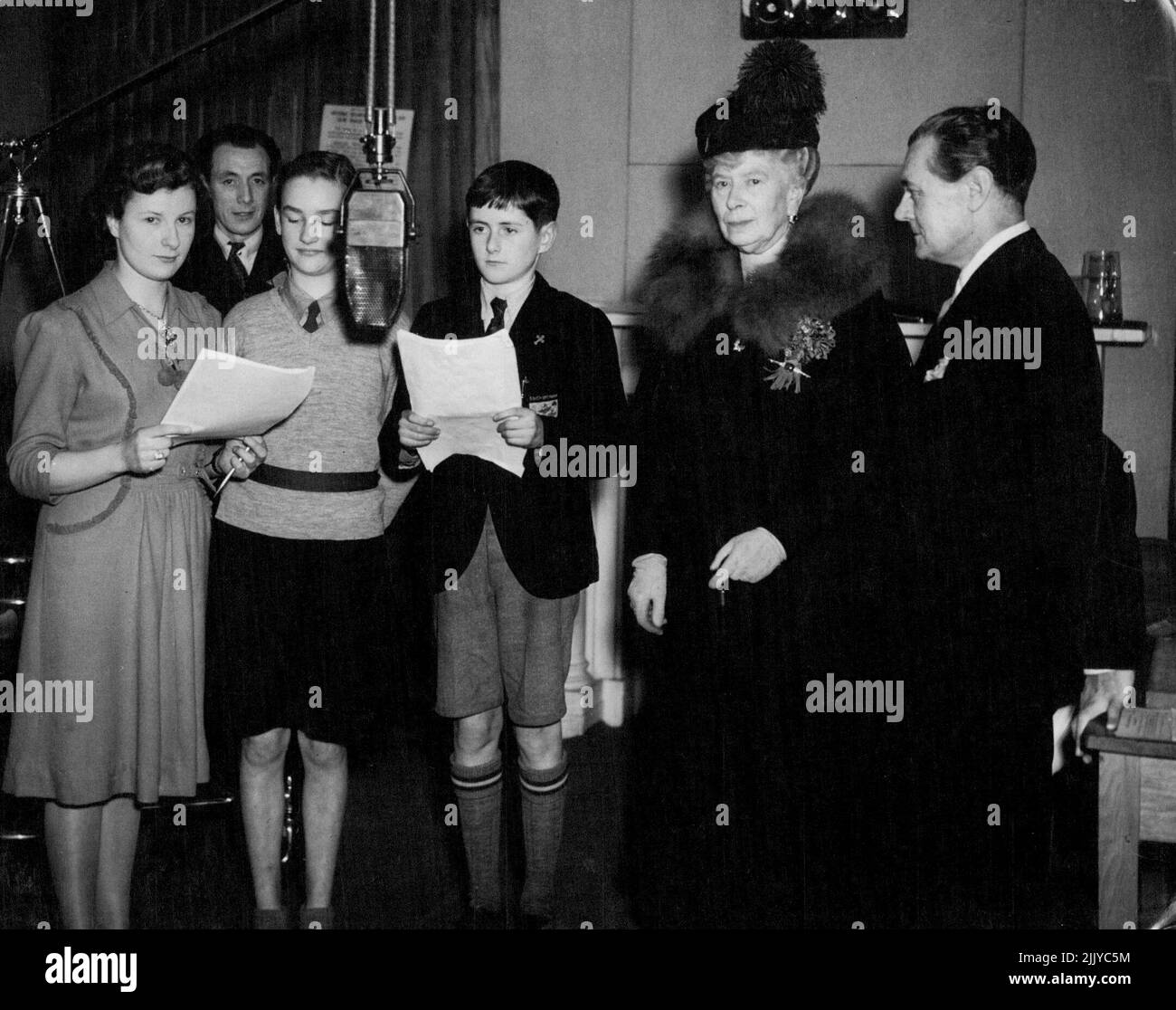 On 25th March, 1943 H.M. Queen Mary paid a visit to Broadcasting House, Bristol, where she saw a schools broadcast transmission, and attended a rehearsal of Ivy Benson's Orchestra, and of the Children's Hour. Queen Mary with Derek McCulloch, at Children's Hour rehearsal of Elizabeth Nesbit's play ' The Railway Children ' - Young players reading from left to right Rosamund Barnes, Jill Andrews, Peter Pullen - with Charles Mason in background. August 09, 1943. (Photo by BBC). Stock Photo