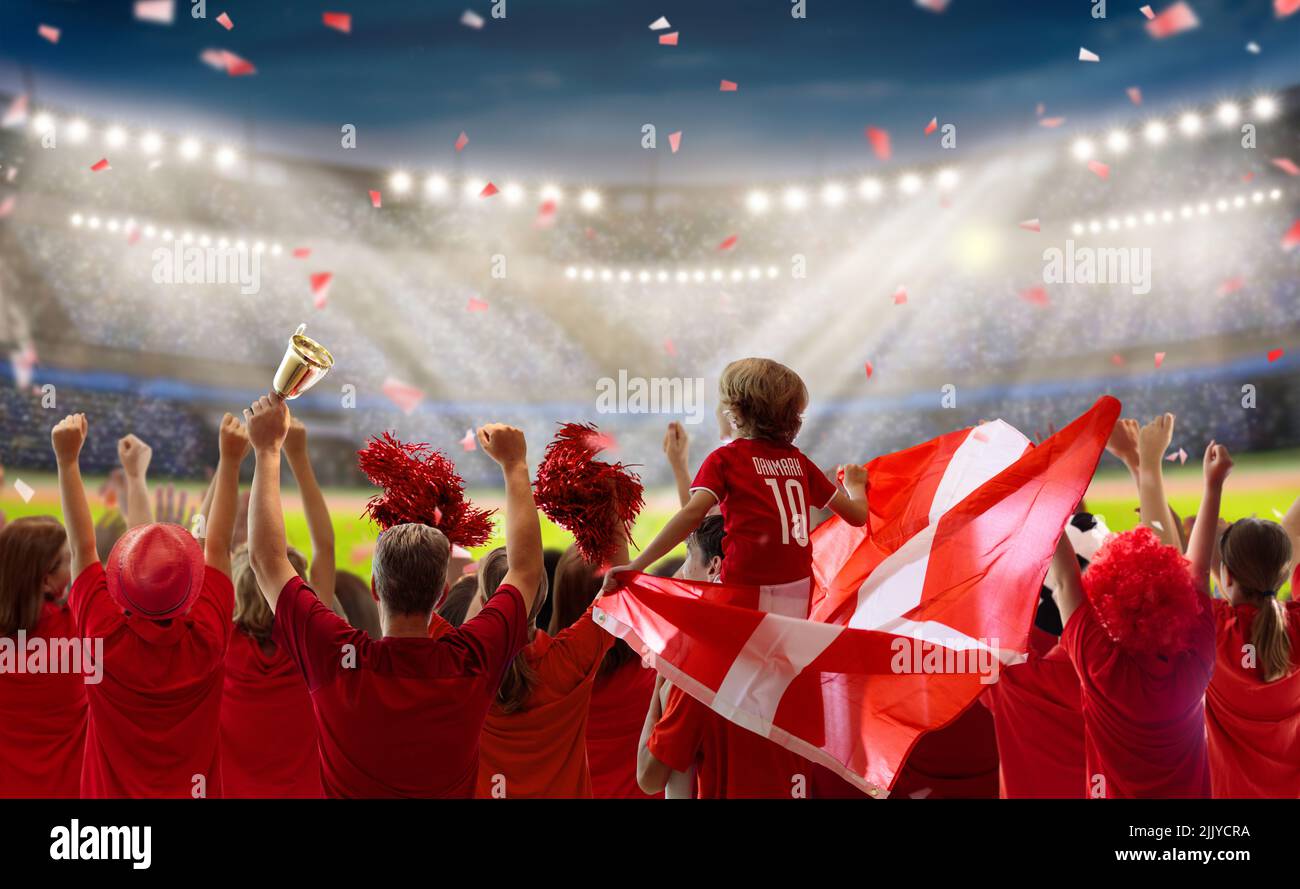 Denmark football supporter on stadium. Danish fans on soccer pitch watching team play. Group of supporters with flag and national jersey Stock Photo