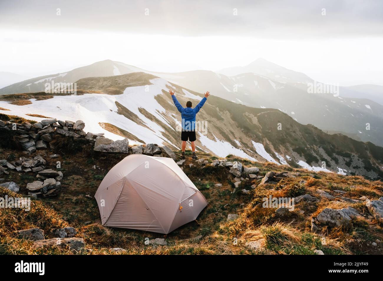 Grey tent and tourist with hand up against the backdrop of an incredible mountain landscape. Amazing evening highland. Tourism concept Stock Photo