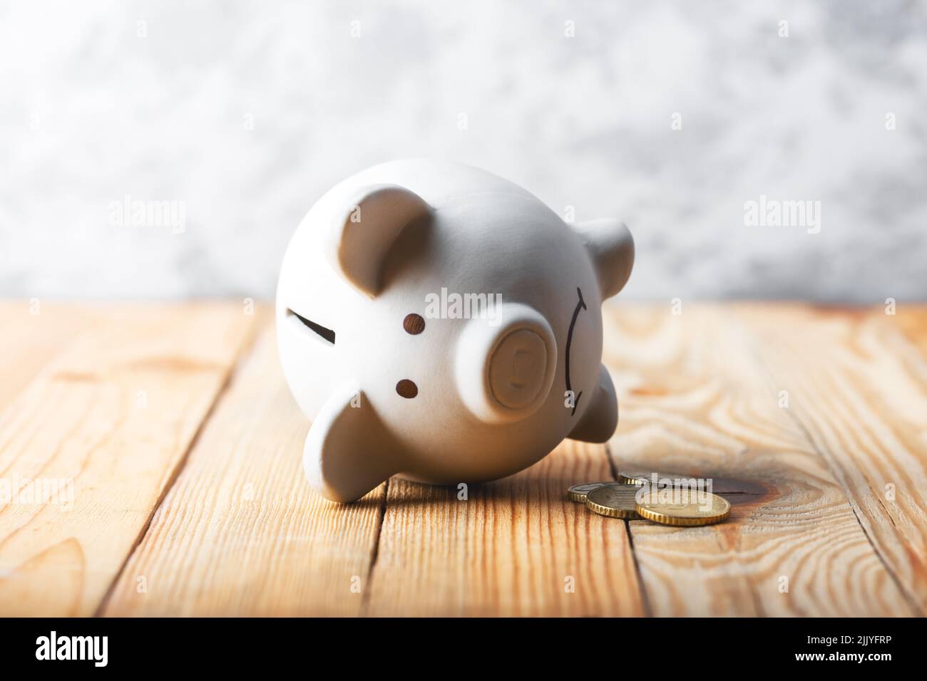 A sad piggy bank behind stack of euro coins symbolizing the fall of monetary assets. White moneybox with golden coins. Crisis concept Stock Photo