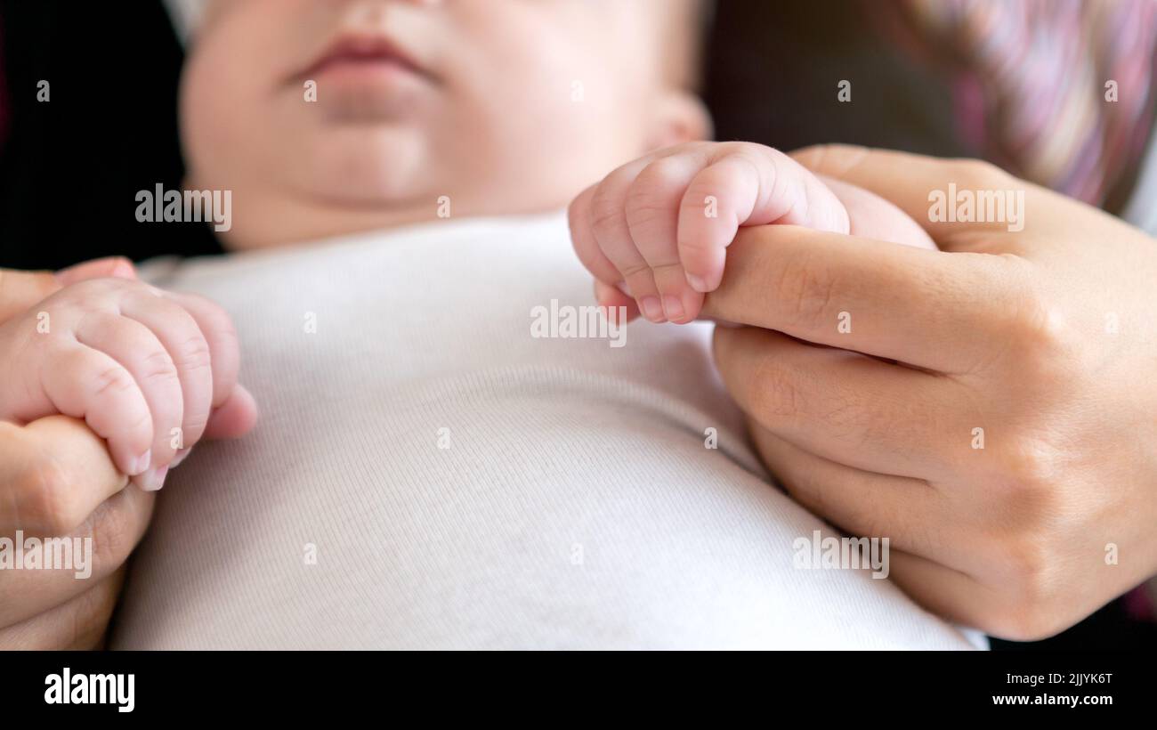 The newborn is holding a finger of mother on a white sheet bed background. A baby boy squeezes a finger of parent. Family and home concept Stock Photo