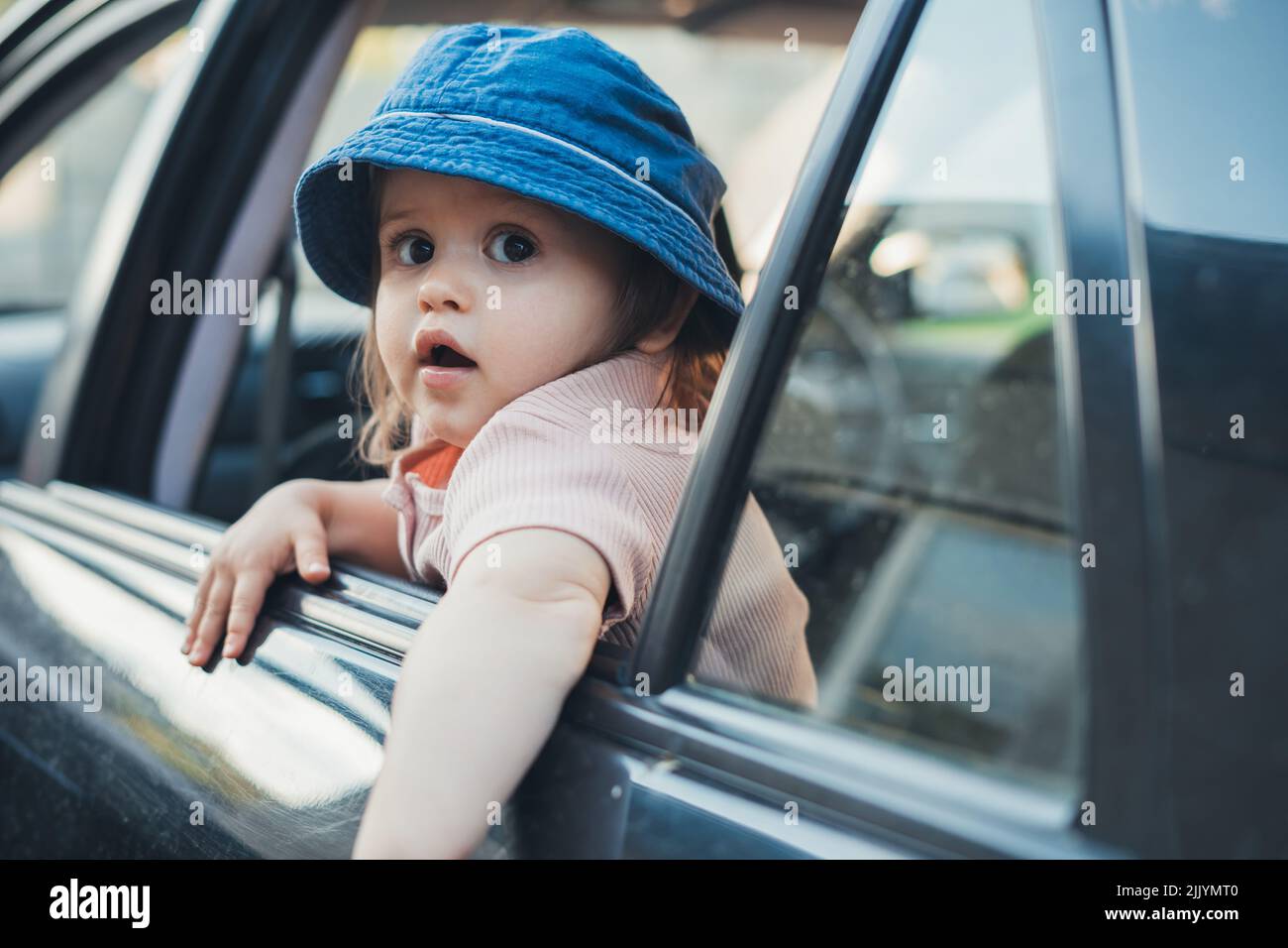 Baby girl sticking her head out of a car window, looking back as he and his family go on a journey. Summer road trip. Traveling with children. Stock Photo