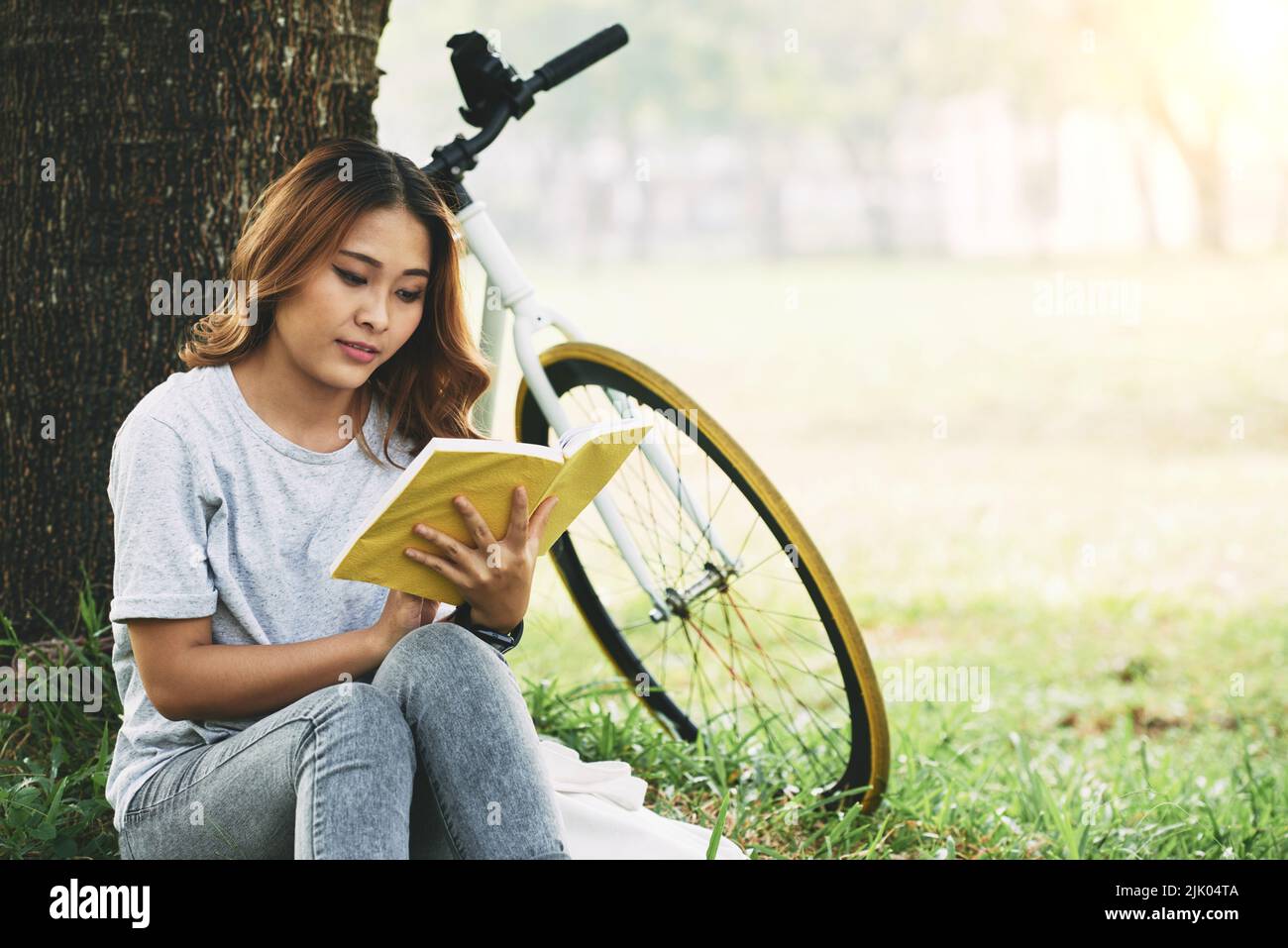 Pretty Vietnmamese young woman sitting under the tree and reading an interesting book Stock Photo