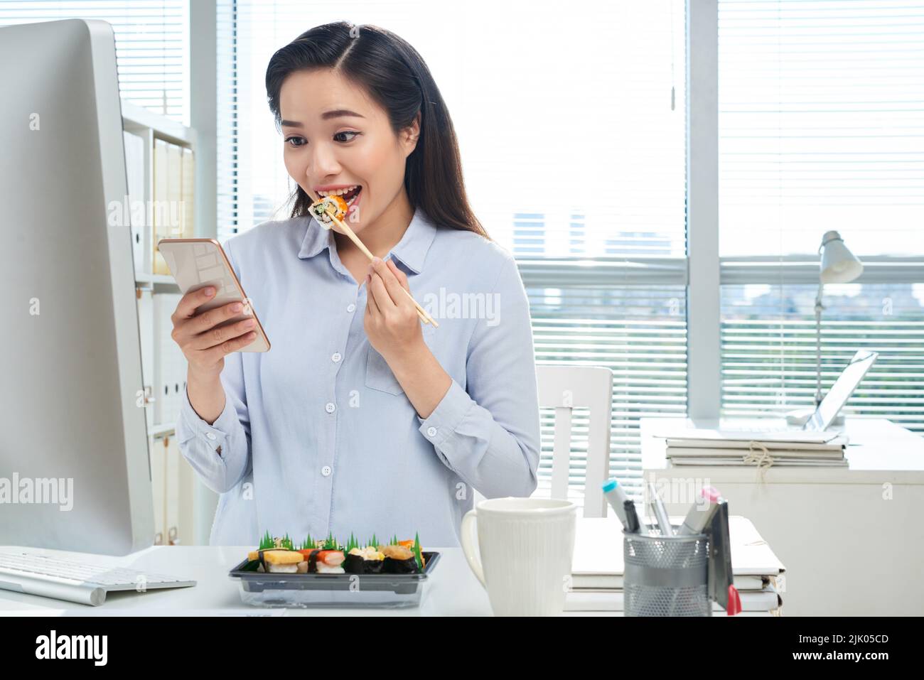 Cheerful Asian white collar worker texting with friend on smartphone while enjoying delicious sushi at workplace, panoramic window on background Stock Photo