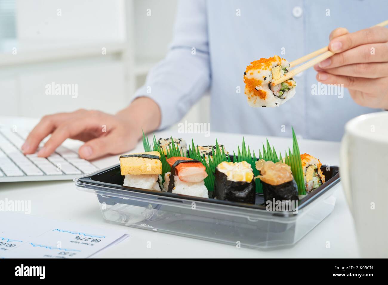 Close-up shot of busy white collar worker sitting in front of computer and eating sushi with help of chopsticks Stock Photo