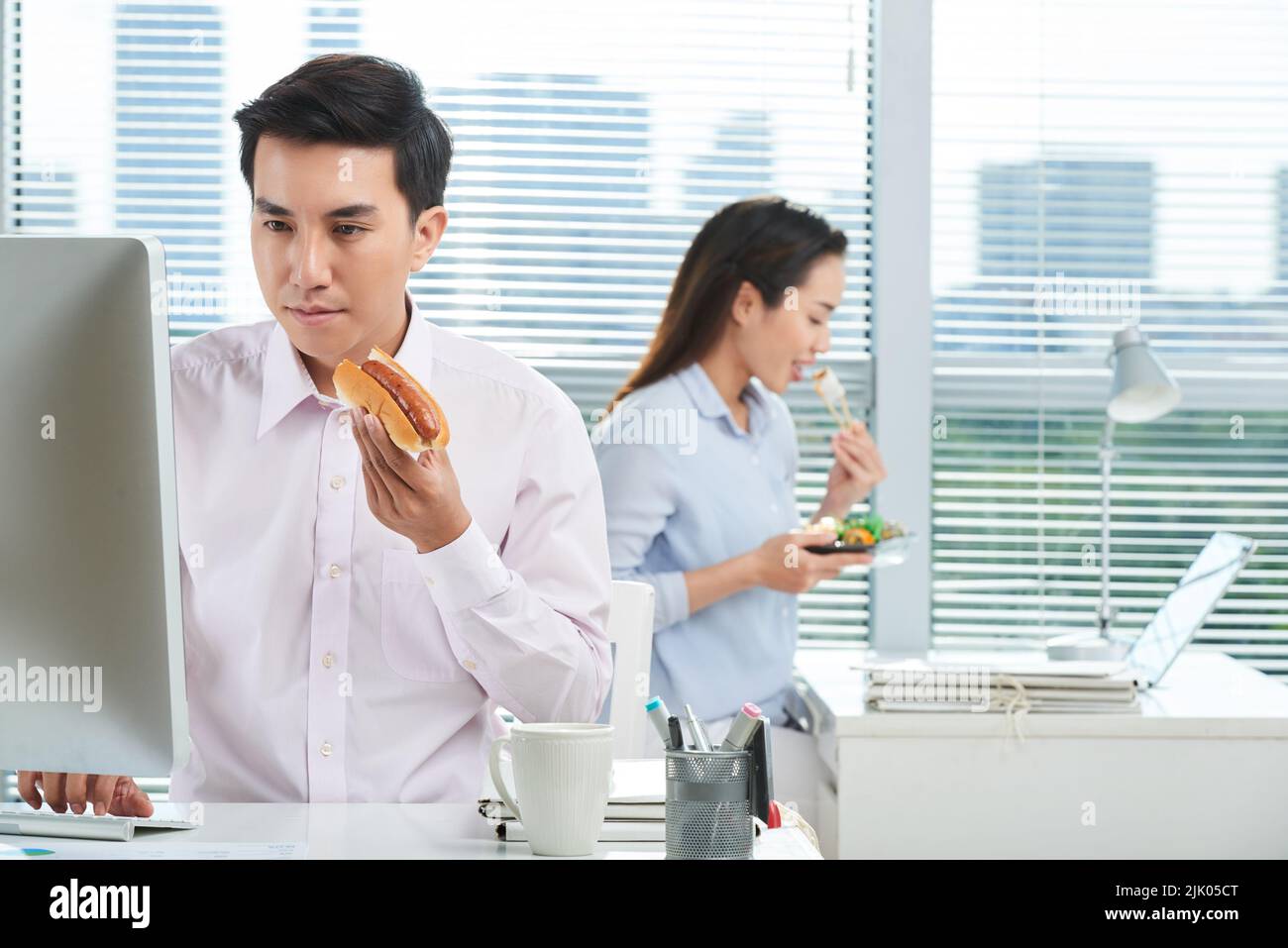 Busy open plan office: concentrated Asian manager wearing white shirt working on computer and eating hot dog while his pretty colleague sitting at workplace and enjoying sushi Stock Photo