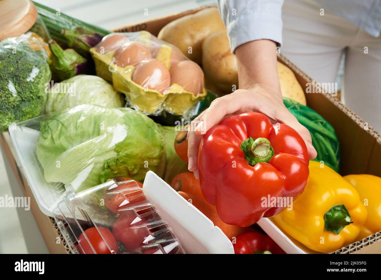 Close-up shot of unrecognizable woman holding bell pepper in hand while checking her online grocery order, cardboard box with vegetables on background Stock Photo