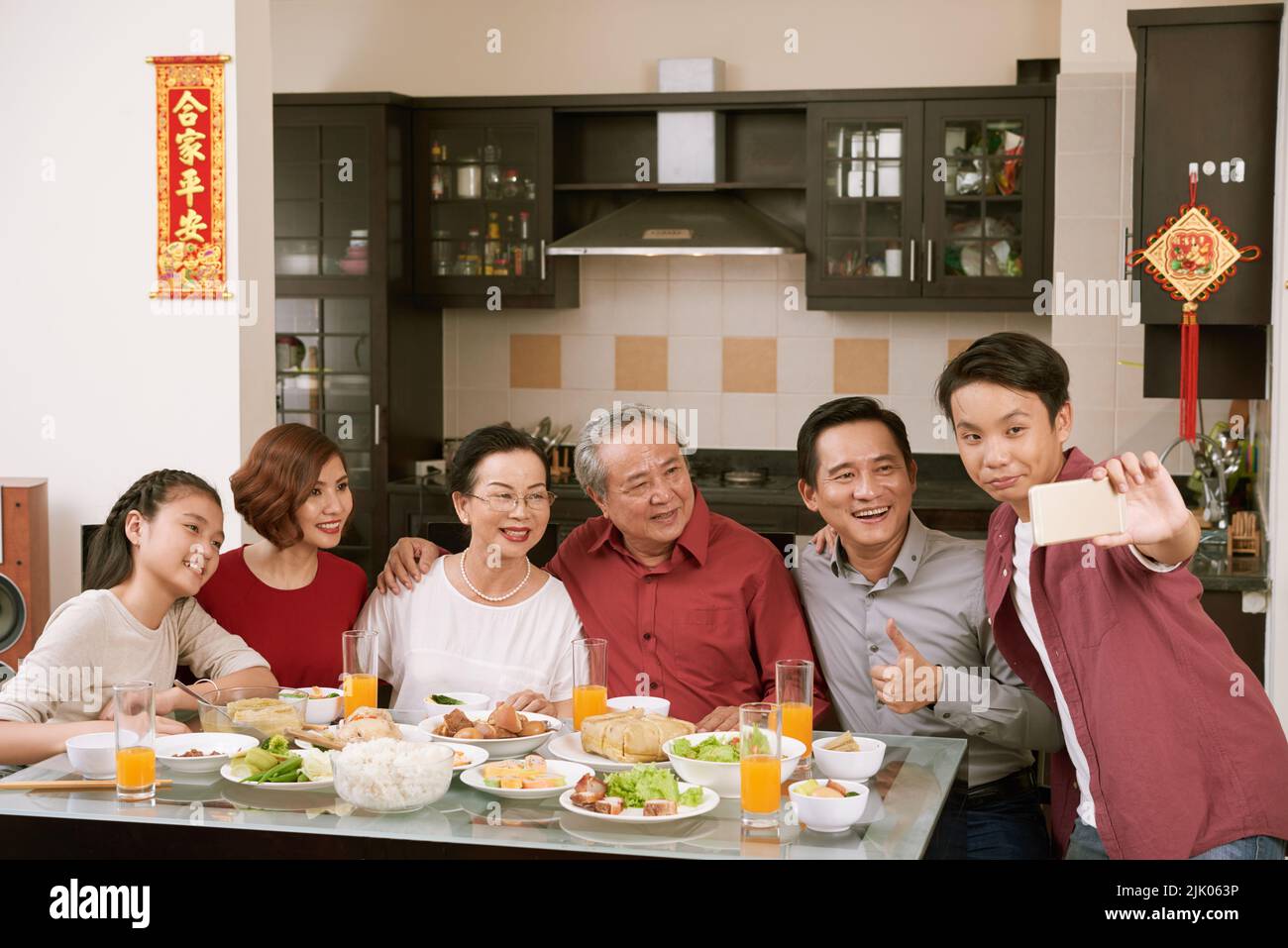 Vietnamese teenage boy taking selfie with his family at Tet celebration dinner. Scrolls wishing luck and wealth in the background Stock Photo