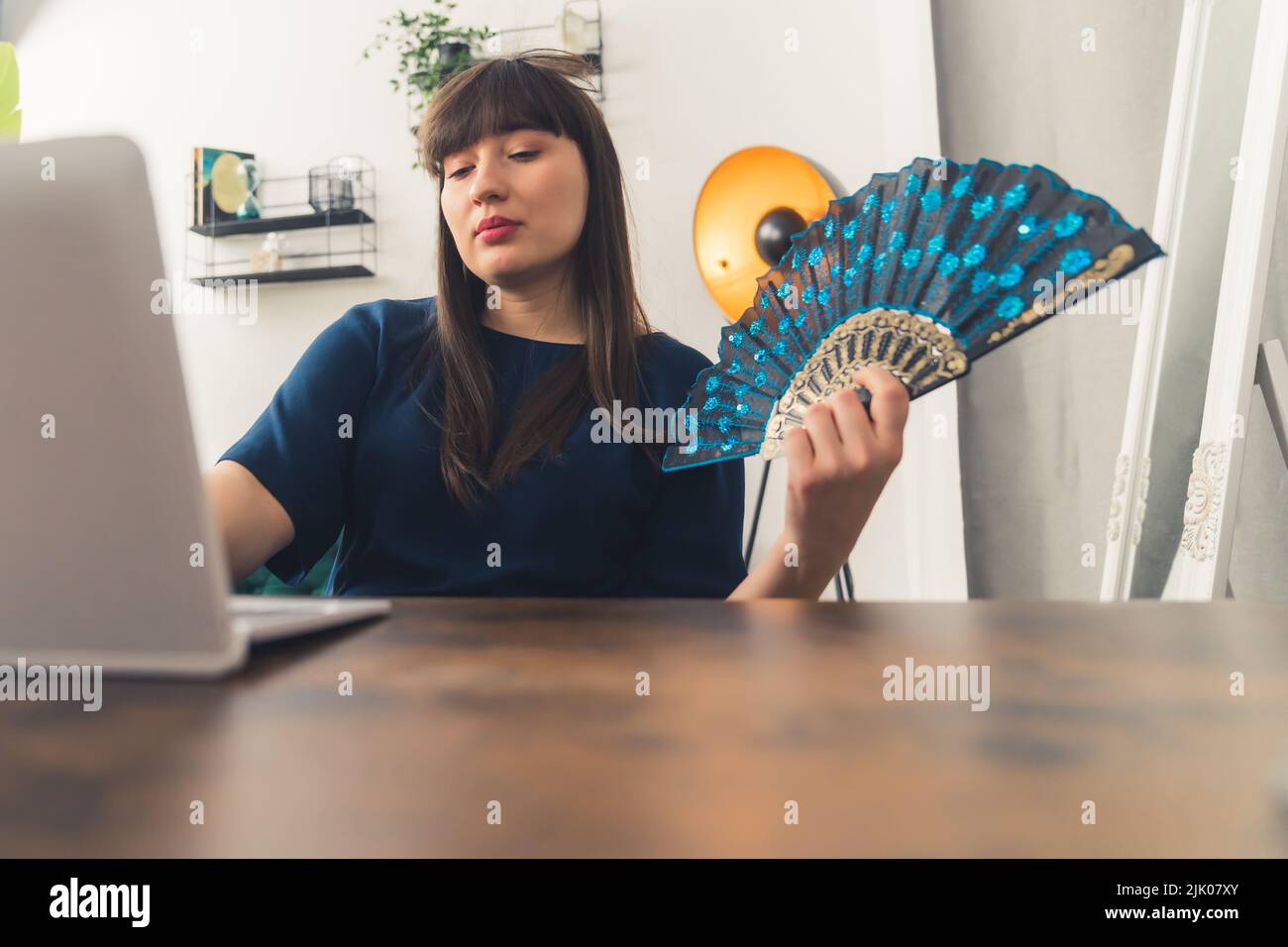 Young brunette female freelancer sits at a table behind her laptop at home and works remotely while using a hand fan because of the summer heat. High-quality photo Stock Photo