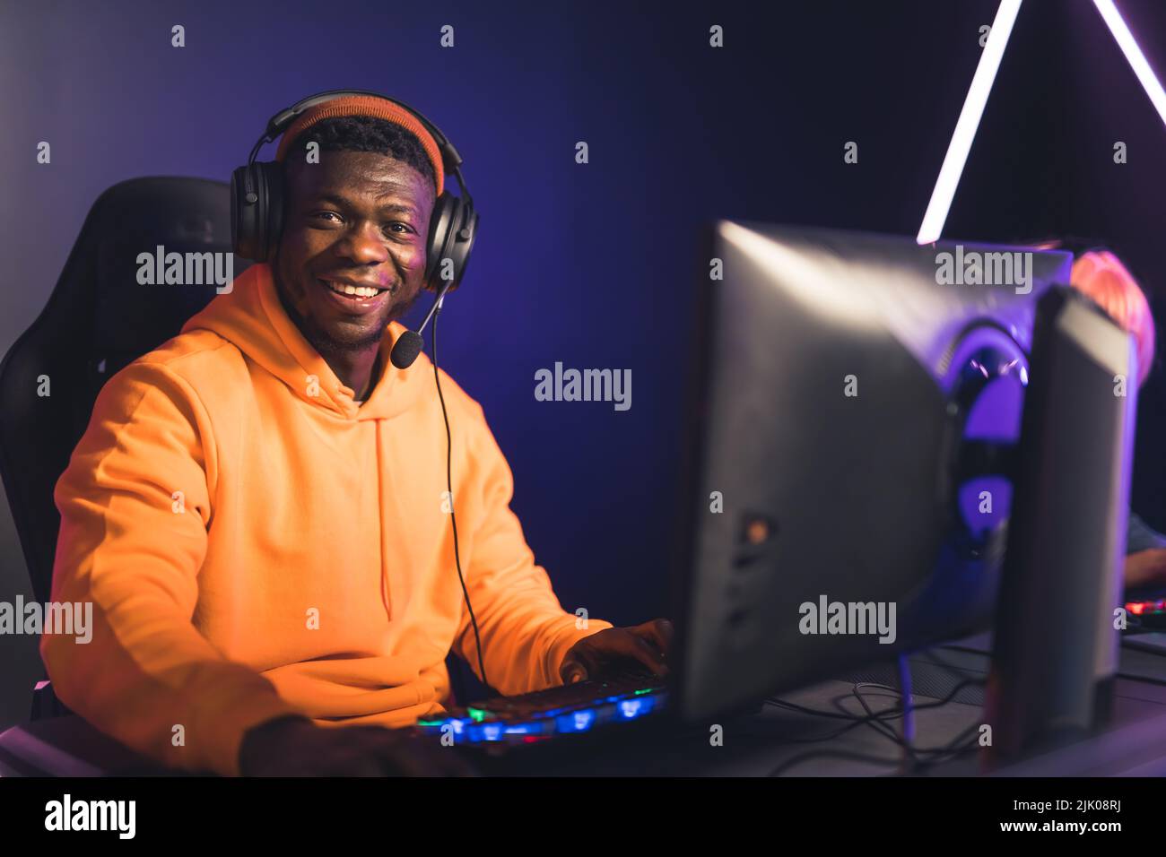 Teenage black guy wearing orange hoodie and headset sitting in gaming chair in front of professional computer smiling into the camera. High quality photo Stock Photo