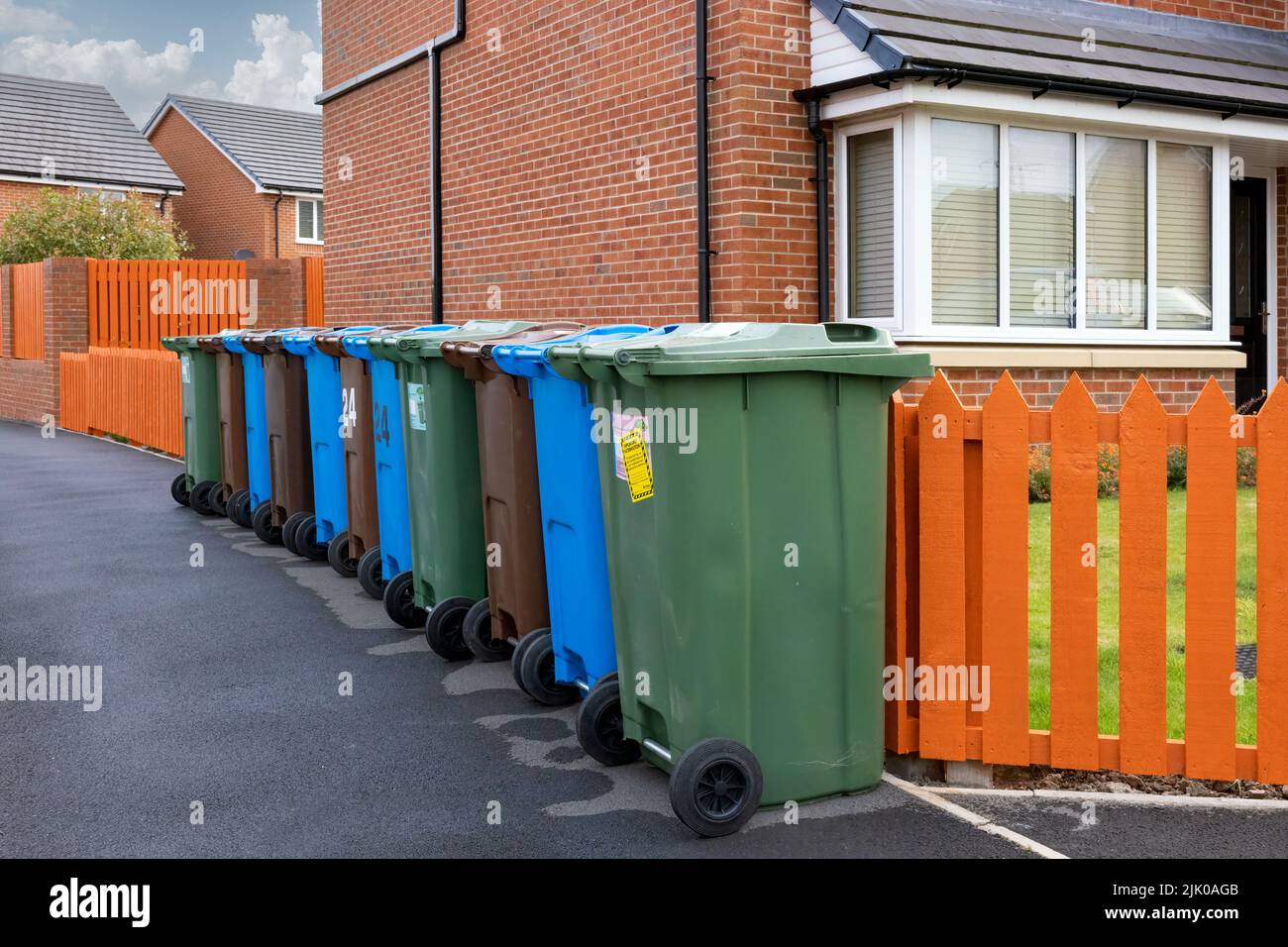 A row of coloured wheelie bins for recycling plastic, paper and garden waste, alongside an orange fence and outside a modern house. Stock Photo