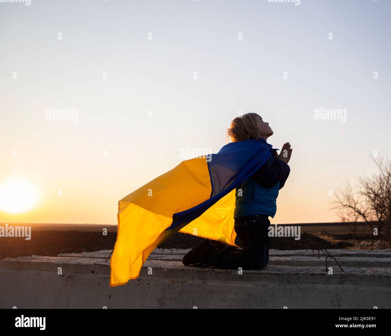 silhouette of boy on his knees, behind child's back flag of Ukraine flutters in wind. hands folded in prayer, appeal to God. Children against war. Ukr Stock Photo