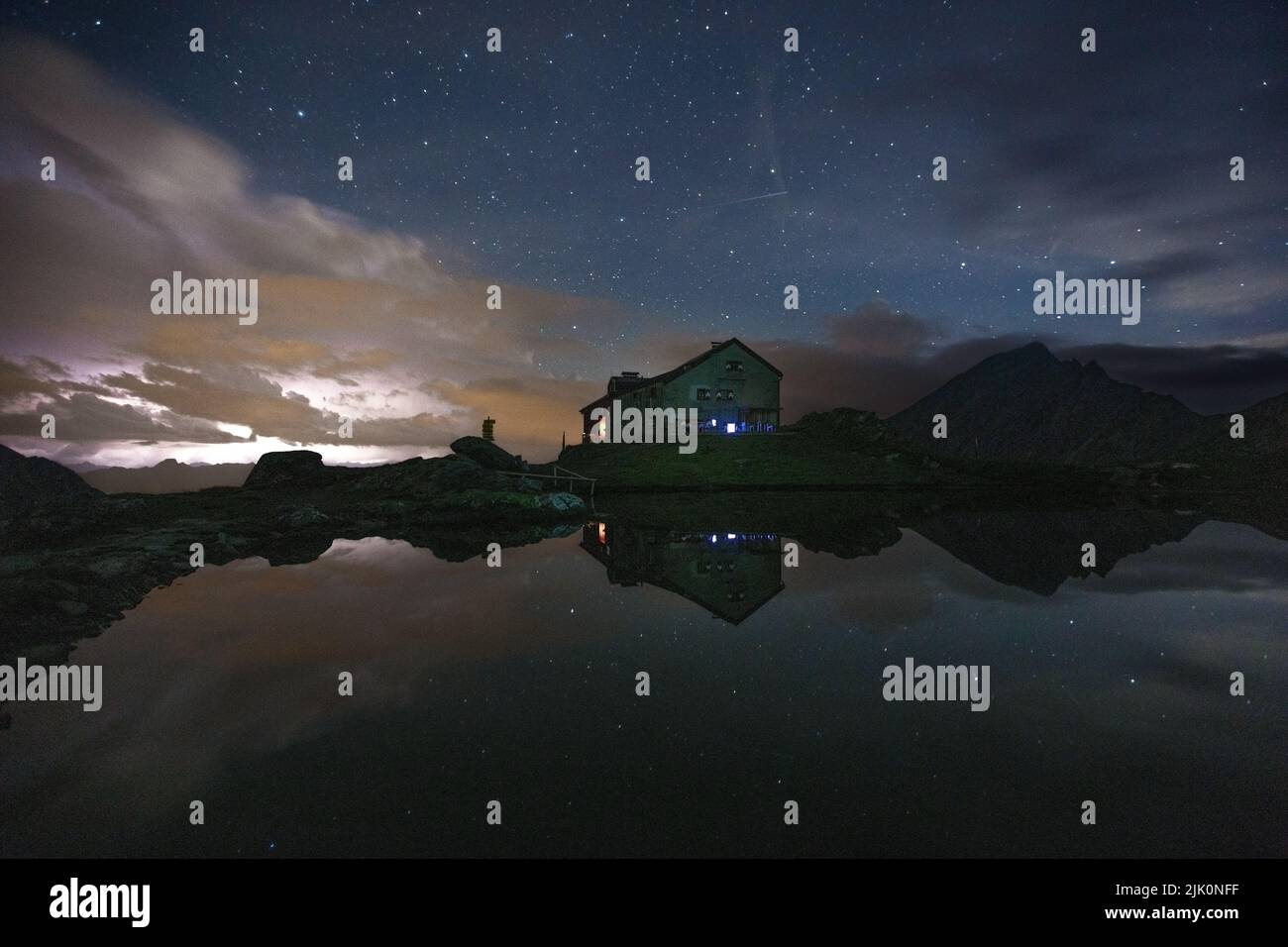 Night at the Sudetendeutsche Hütte alpine refuge. Thunderstorm, lightnings in the background, starry sky reflecting on the lake water. Austrian Alps. Stock Photo