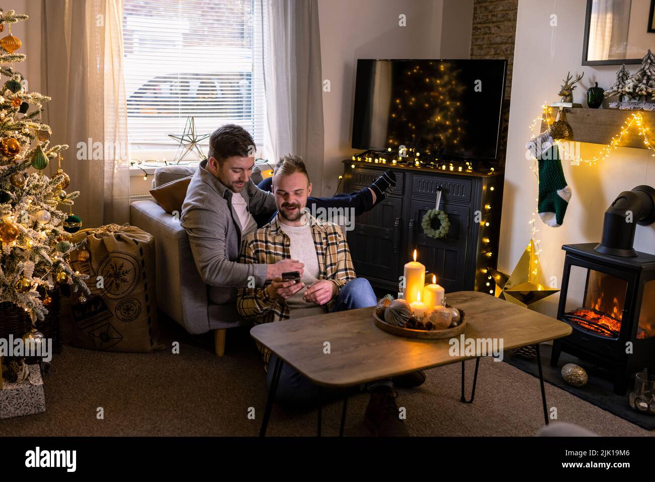 A front-view shot of an LGBTQI same sex male couple sitting in their living room relaxing, one man is showing his partner his smart phone, the house i Stock Photo