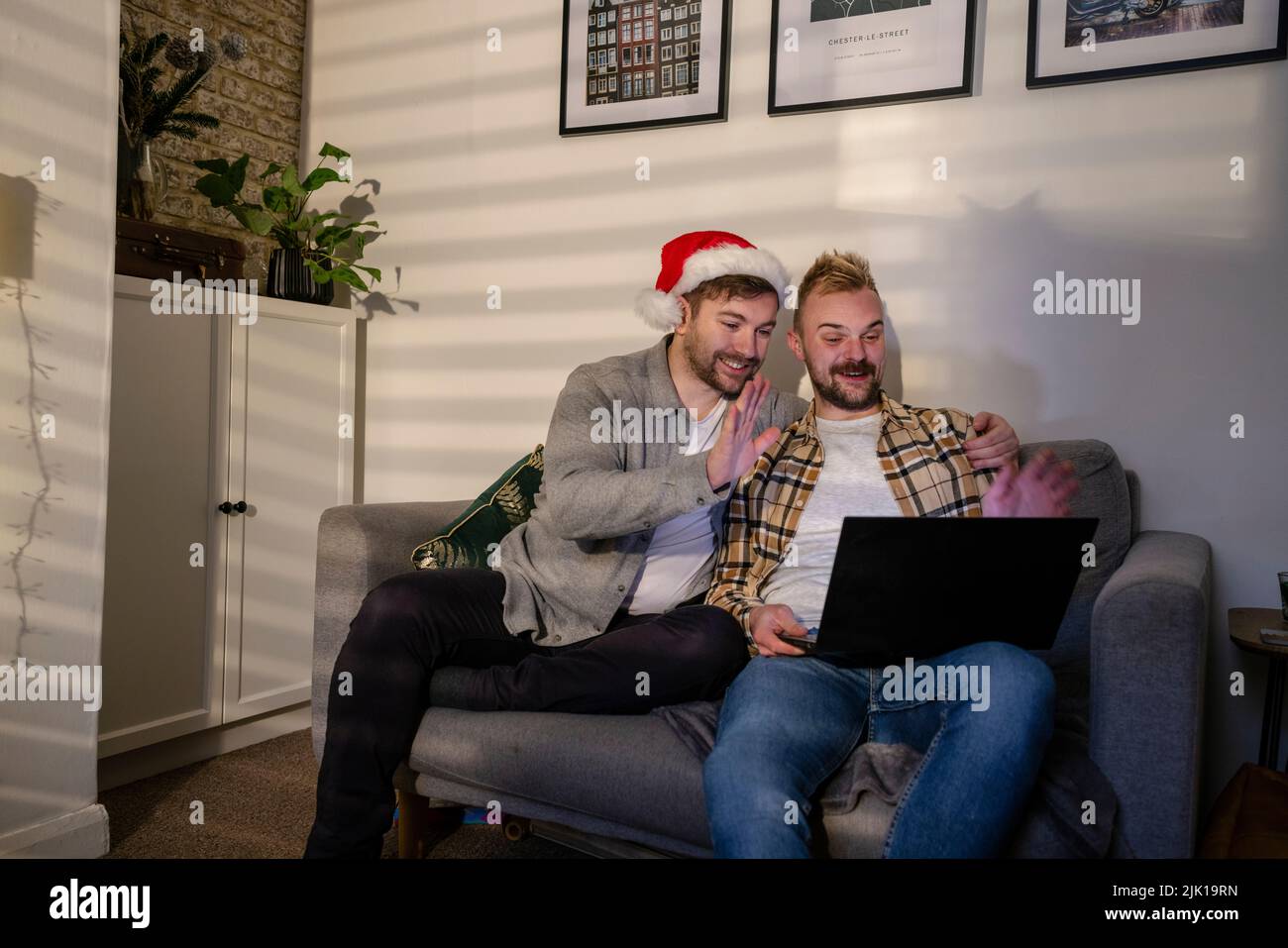 An LGBTQI same sex male couple sitting on the sofa in their living room relaxing, they are using a laptop and waving at their friend on a video call. Stock Photo