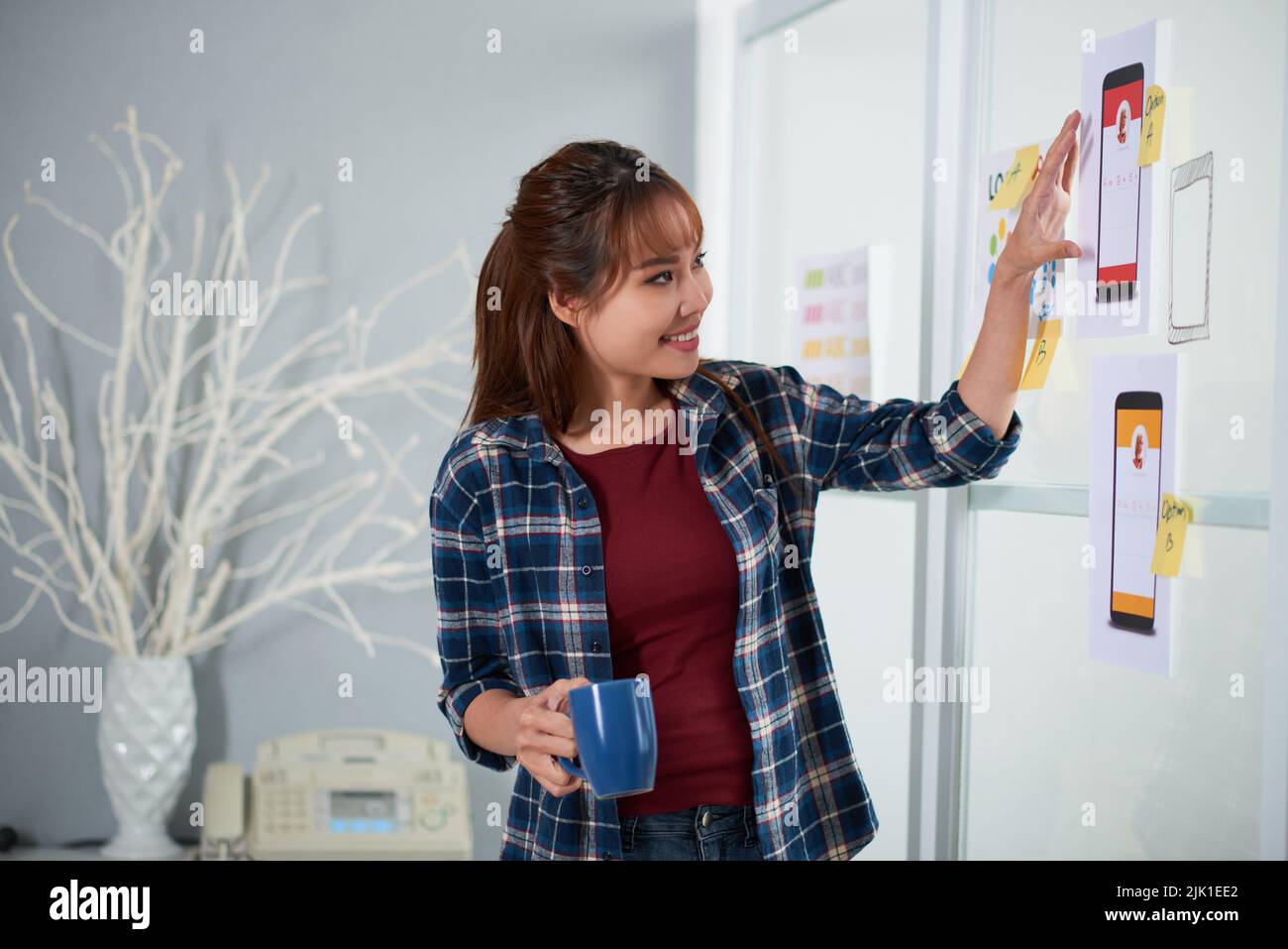 Cheerful Asian designer standing at whiteboard with cup of coffee in hand and brainstorming on start-up project, interior of stylish studio on background Stock Photo