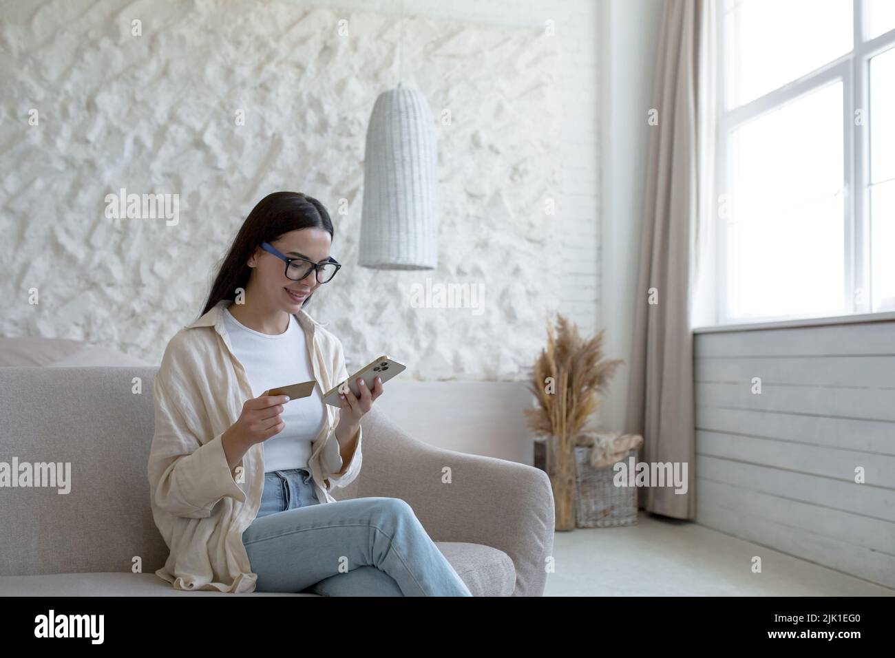 Beautiful young woman at home in glasses sitting on the sofa using a mobile phone smartphone for online shopping in a store, holding a bank credit card in her hands Stock Photo
