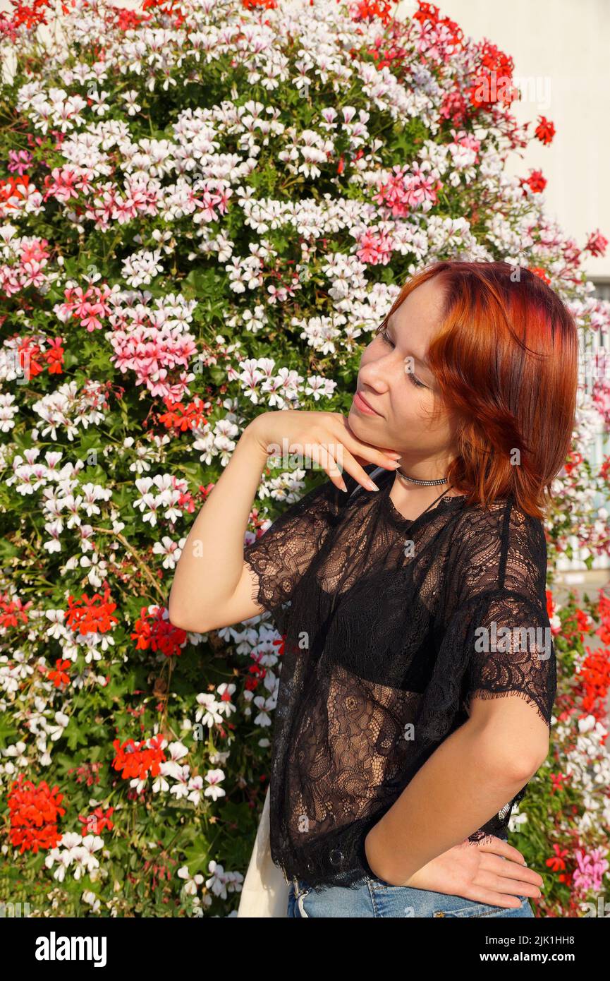 portrait of a smiling red-haired teenage girl with closed eyes on a floral background in the sunlight. Stock Photo
