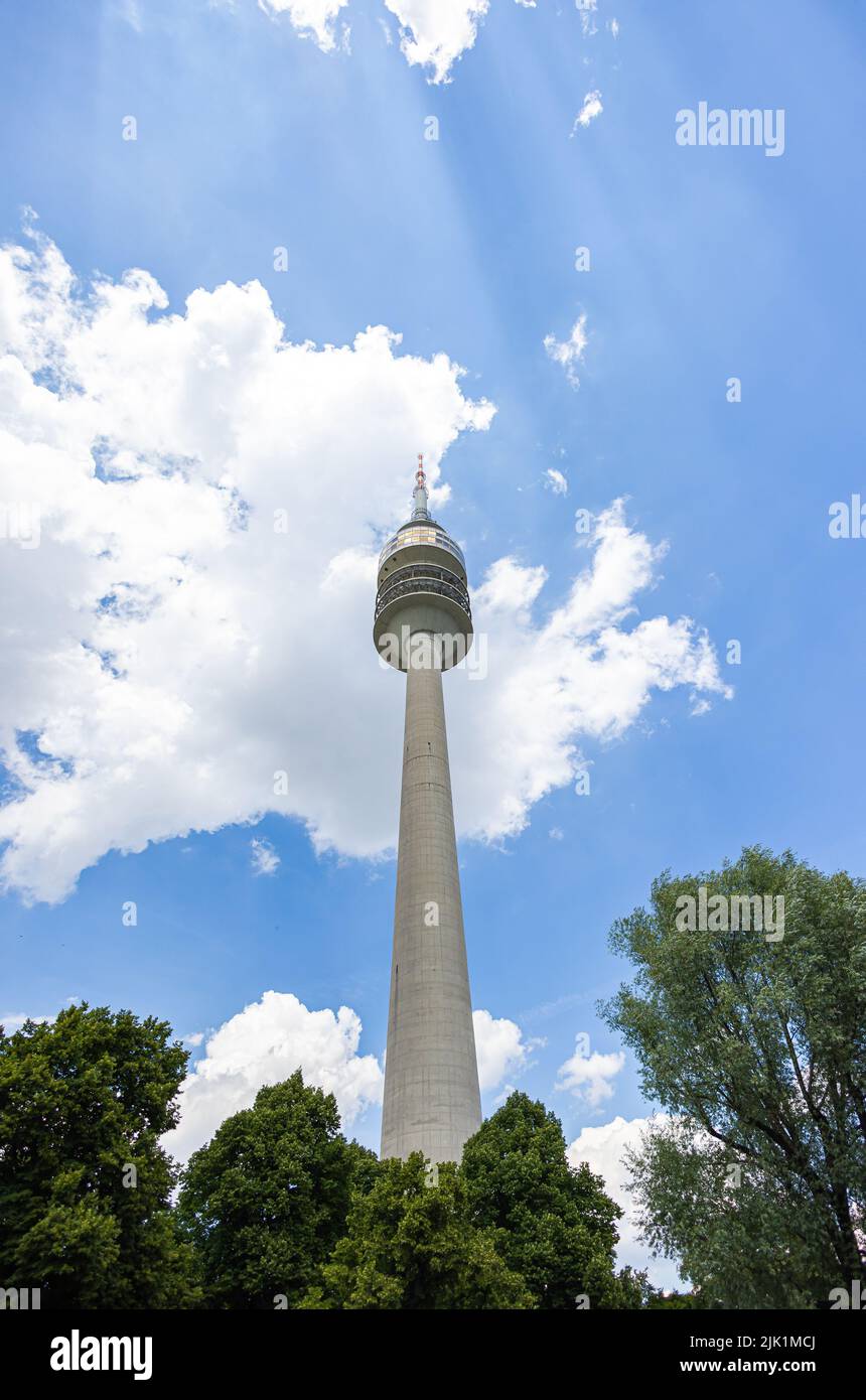 Munich, Germany - July 6, 2022: The Olympiaturm (Olympia tower) at Olympia park. Looking up through the trees of the park. Blue sky and white cloud be Stock Photo
