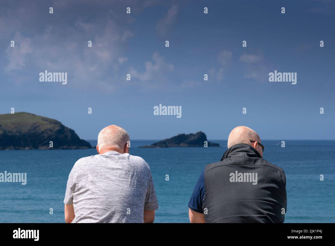 Two male mature holidaymakers looking out to the rocky island called The Goose in Fistral Bay in Newquay in Cornwall in the UK. Stock Photo