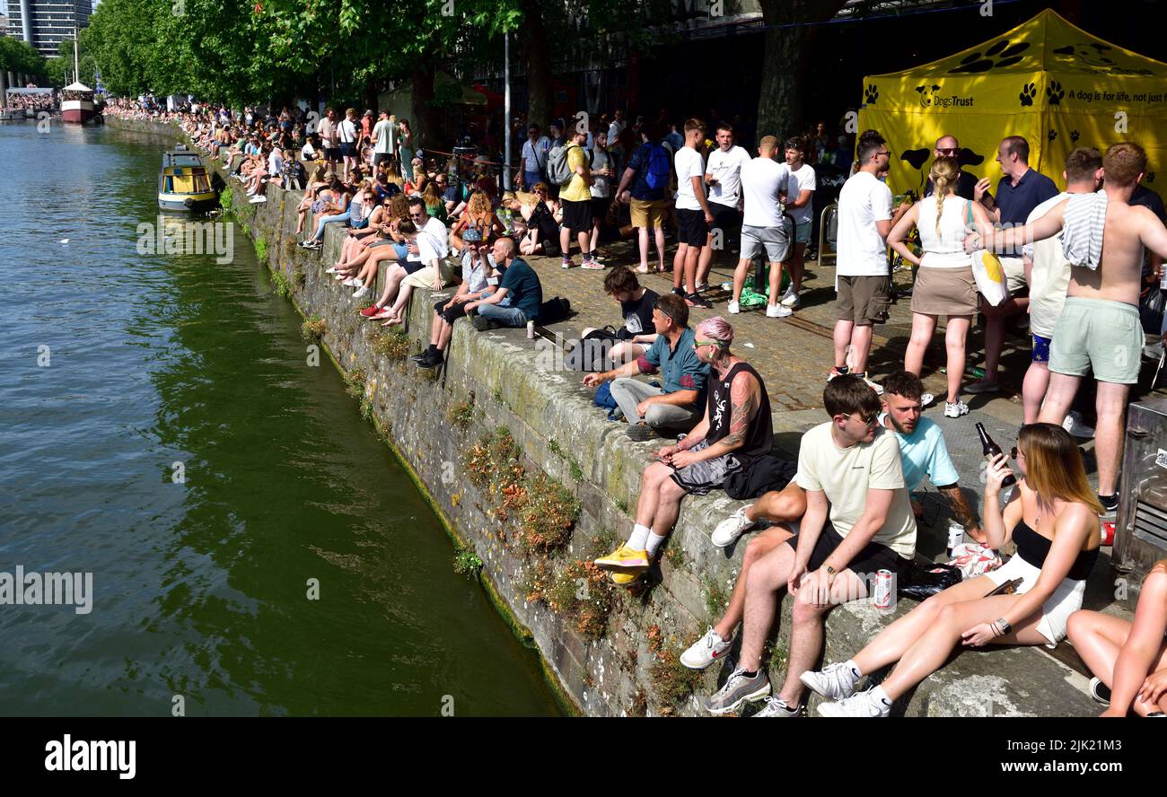 People enjoying the sun sitting along Bristol narrow quay wall on a sunny hot day during harbour festival, UK Stock Photo