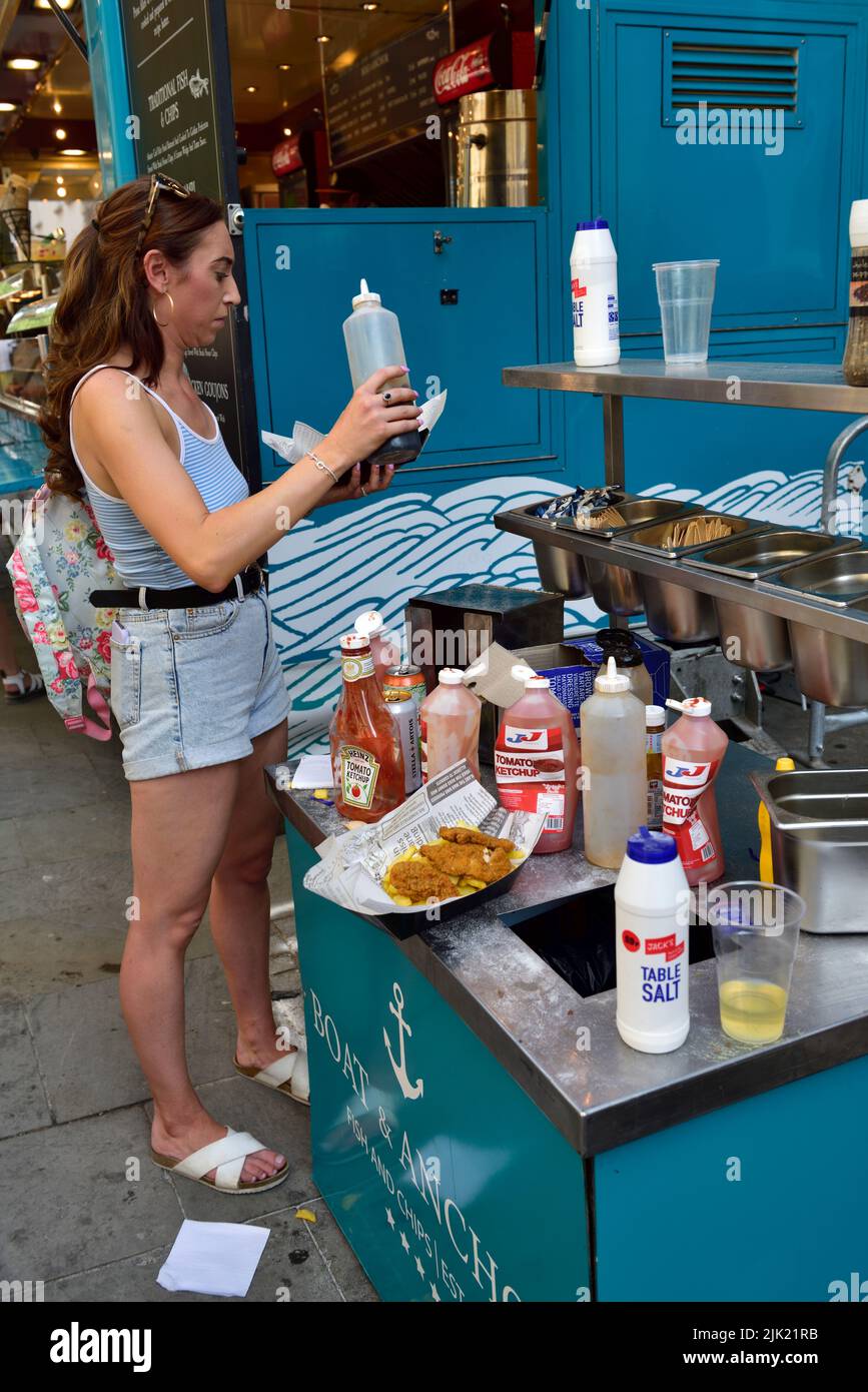 Woman putting vinegar on order of fish and chips next to a condiments selection at festival Stock Photo