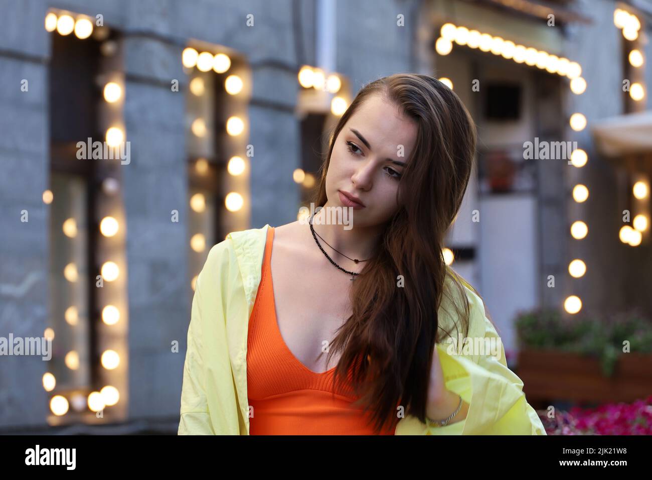 Portrait of pretty young girl with long hair and perfect makeup wearing orange top and yellow shirt on a street on blurred lights background Stock Photo