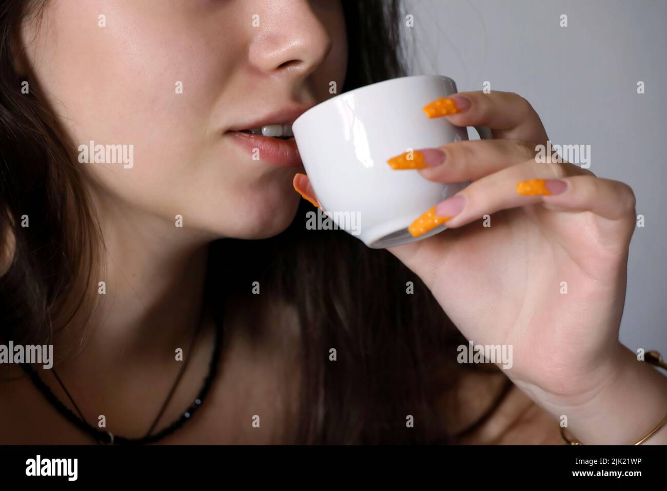 Sensual girl drinking tea or coffee, female lips and white cup in hand with orange acrylic nails close up. Concept of enjoying hot drink Stock Photo