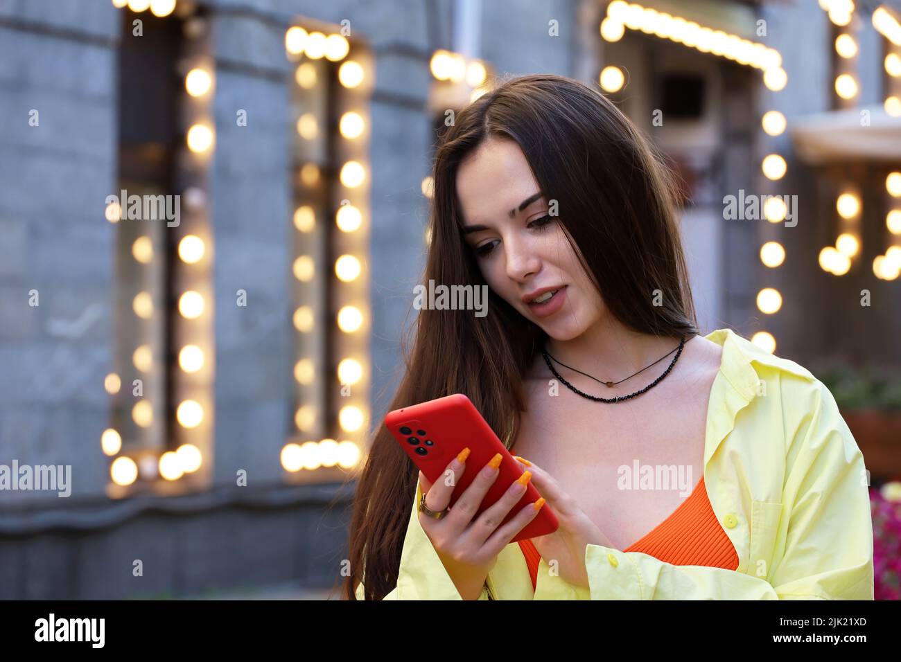 Smiling young girl with long hair and perfect makeup using smartphone on a street on festive lights background. Female beauty, online communication Stock Photo