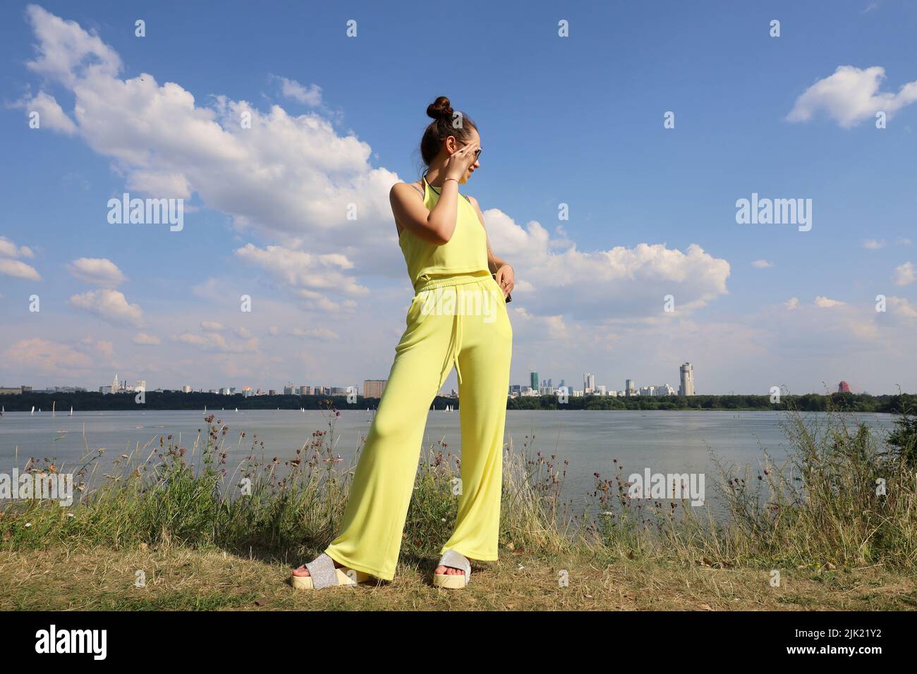 Happy slim girl in sunglasses and yellow summer suit standing on a beach on background of city buildings. Females fashion and beauty Stock Photo