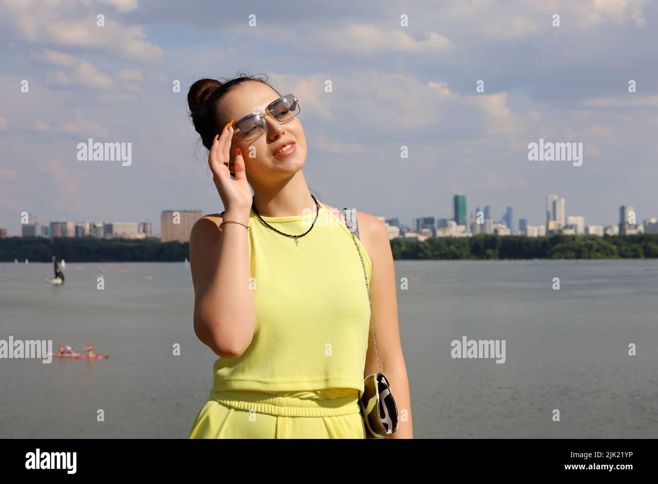 Portrait of sensual girl in sunglasses and yellow summer suit standing on a beach on background of city buildings. Females fashion and beauty Stock Photo