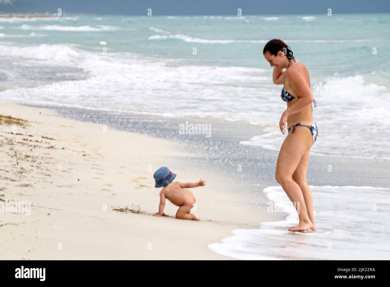 Miami Beach Florida,Atlantic Ocean Shore shoreline coast coastline seashore,baby babies child wearing hat,crawling near surf naked mother watching Stock Photo