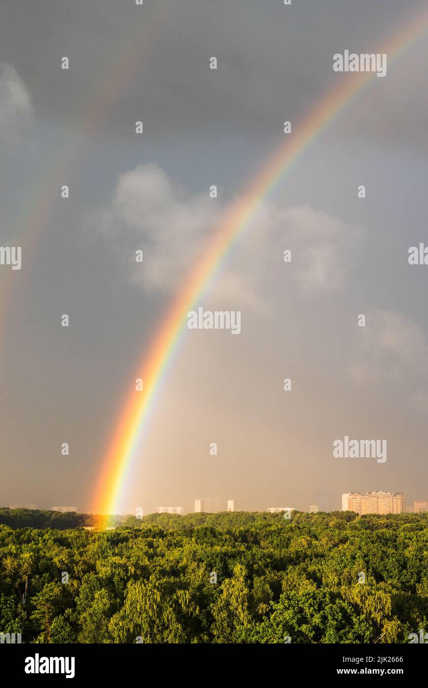 bright rainbow in gray blue sky over sunlit city park before thunderstorm on sunny summer day Stock Photo