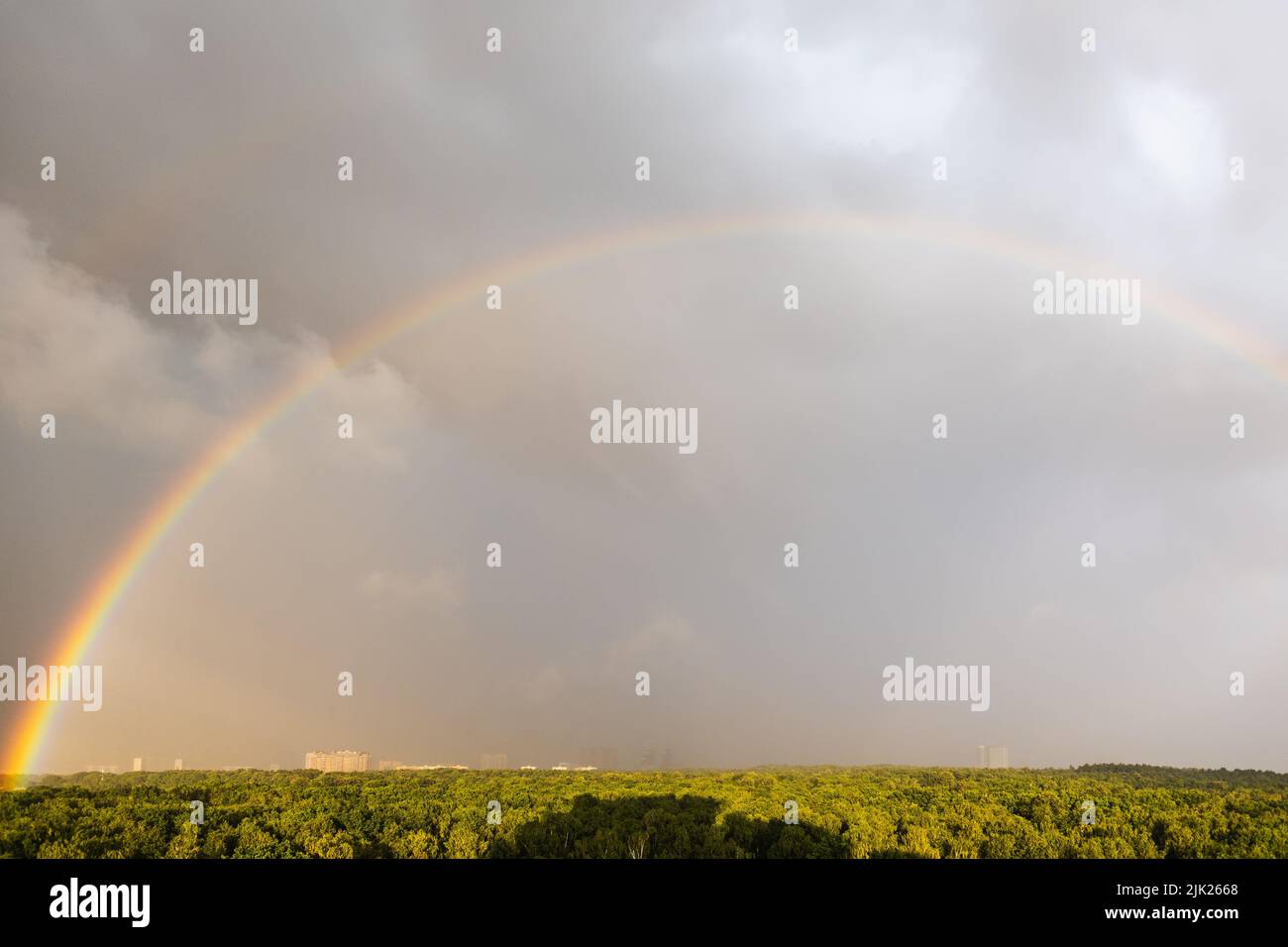 large rainbow in gray blue sky over sunlit city park before thunderstorm on sunny summer day Stock Photo