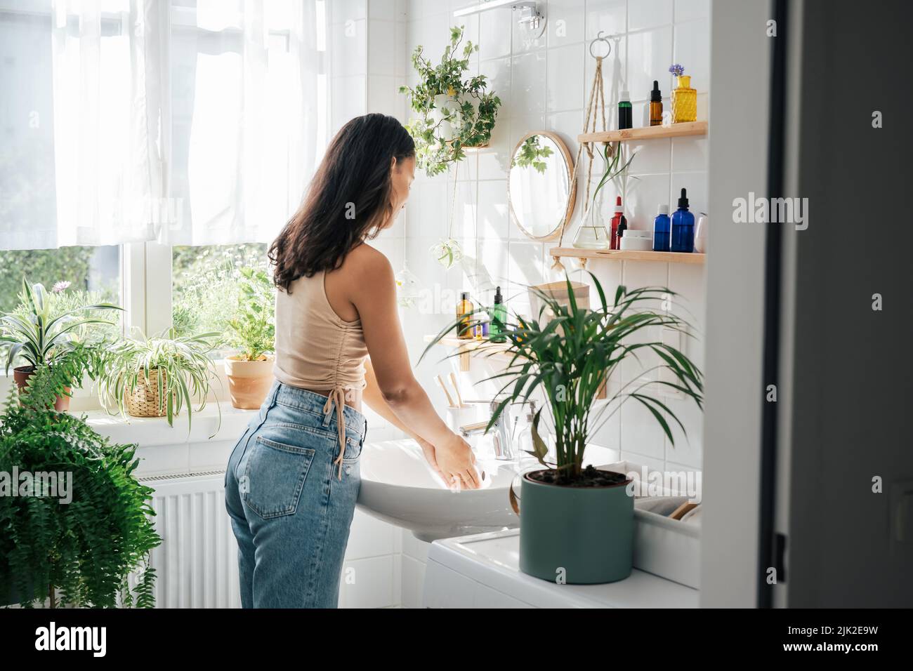 Young beautiful woman with dark skin washing hands in light bathroom with many green plants. Biophilic design of interior. Natural cosmetics, self care and wellness concept Stock Photo
