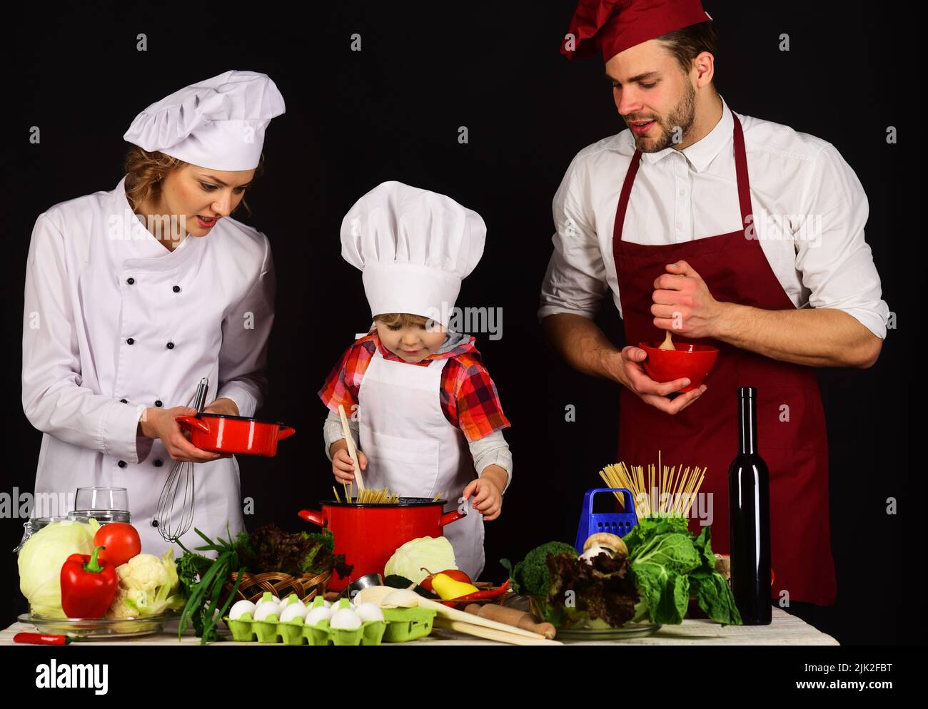 Child with parents cooking at kitchen. Happy family in chef uniform preparing dinner together. Stock Photo