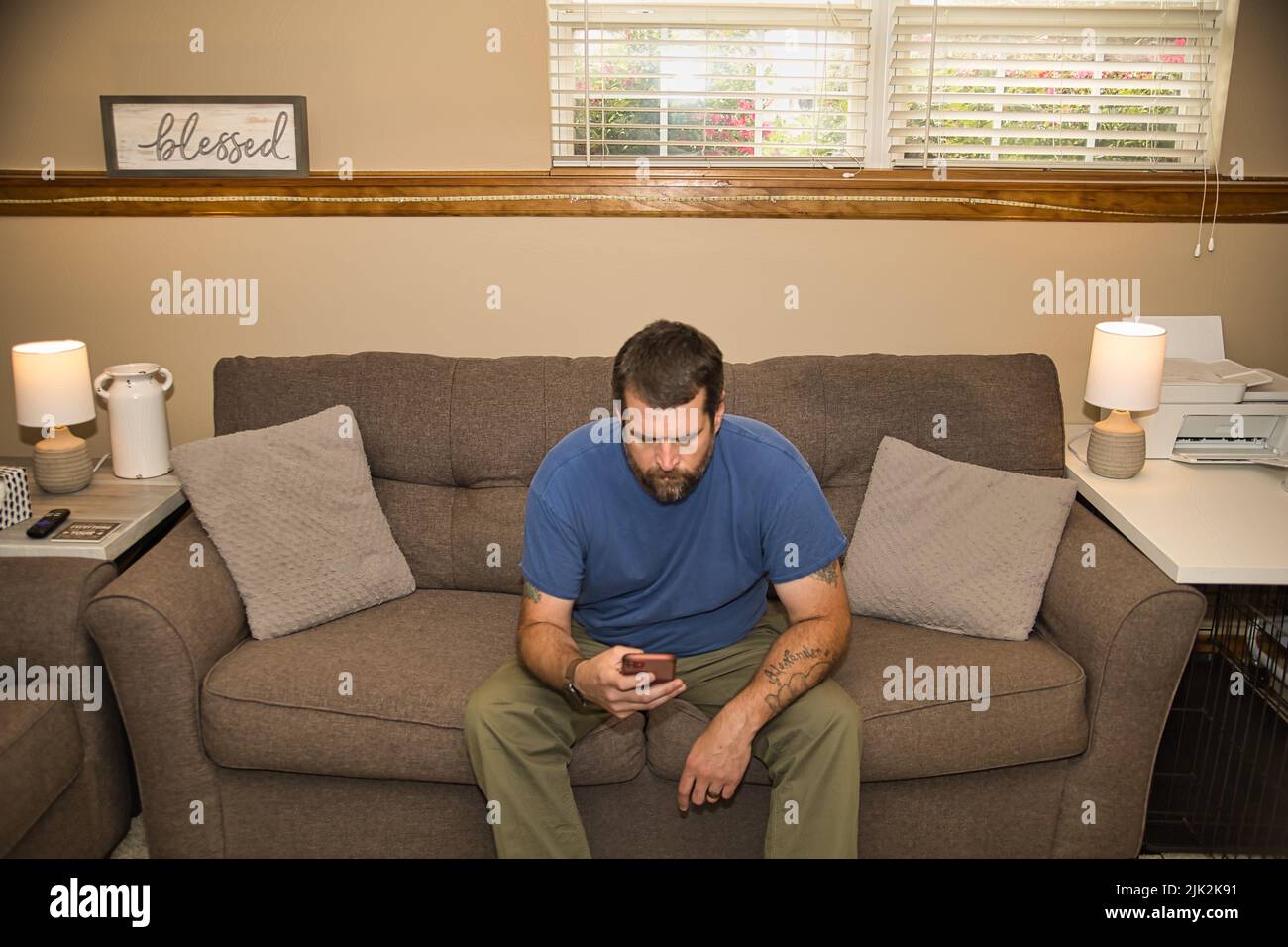 This dark haired bearded man is looking at his phone while sitting on the gray couch. Stock Photo