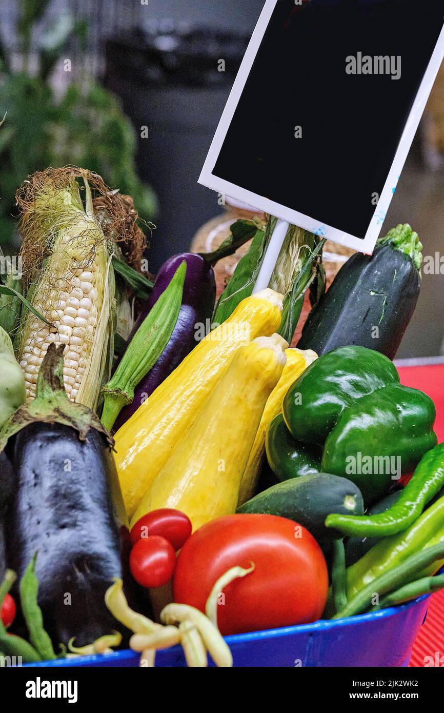 A basket of farm fresh tomatoes, bell peppers, eggplant, and corn.  A small blank sign with black field is in place for additional text. Stock Photo