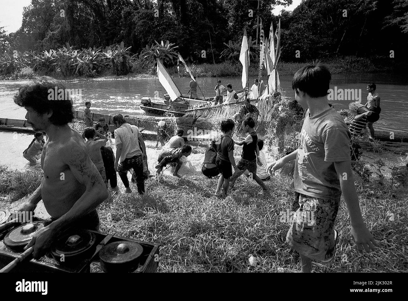 Uluk Palin village, Embaloh Hilir, Kapuas Hulu, West Kalimantan, Indonesia. March 2007. A man unloading a set of gamelan percussive instrument walking pass a crowd of people--Dayak Tamambaloh community--who splash water at each others as a purifying act, in front of a boat that just carried the family of the new traditional chief for a visit to a nearby cemetery where their former chief is buried. This is a part of a series of traditional events held to honour the former traditional chief, who passed away several weeks earlier.--Photographed on black and white film, scanned, digitalized. Stock Photo