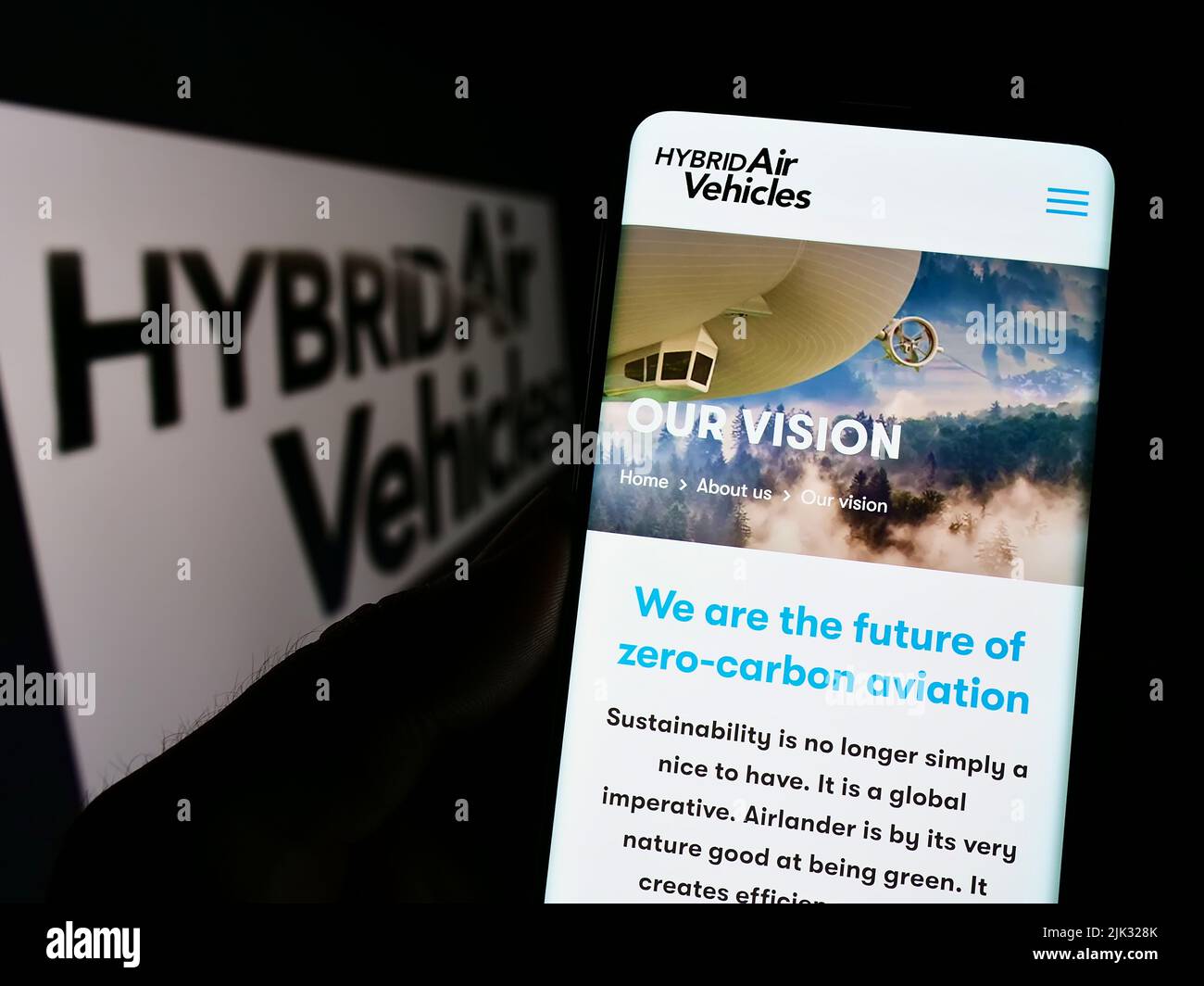 Person holding cellphone with webpage of company Hybrid Air Vehicles Limited (HAV) on screen in front of logo. Focus on center of phone display. Stock Photo