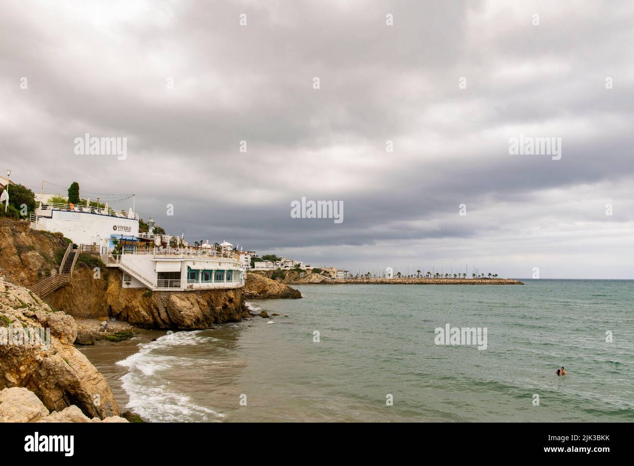 A couple swimming at the mediterranean sea in the rocky coasts near San Sebastian beach at Sitges, Barcelona Province. Sitges port at the bottom Stock Photo