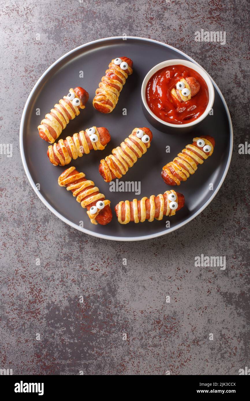 Scary mummy sausages in the dough with funny eyes on the table close-up. Funny decorations. Halloween Food. Vertical top view from above Stock Photo