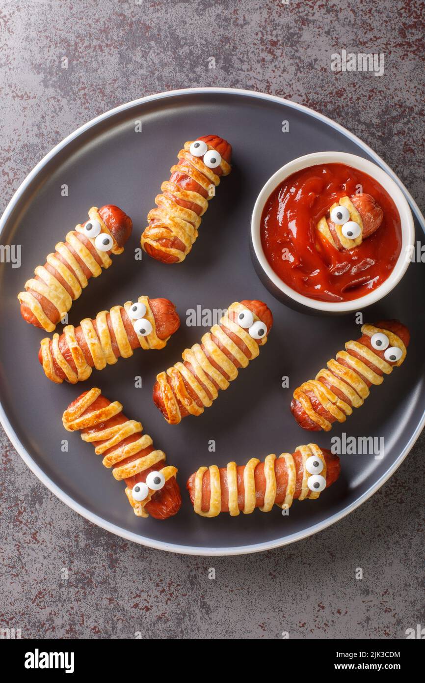 Mummy sausage in dough scary Halloween holiday food with funny eyes and sauce on a plate on the table. Vertical top view from above Stock Photo